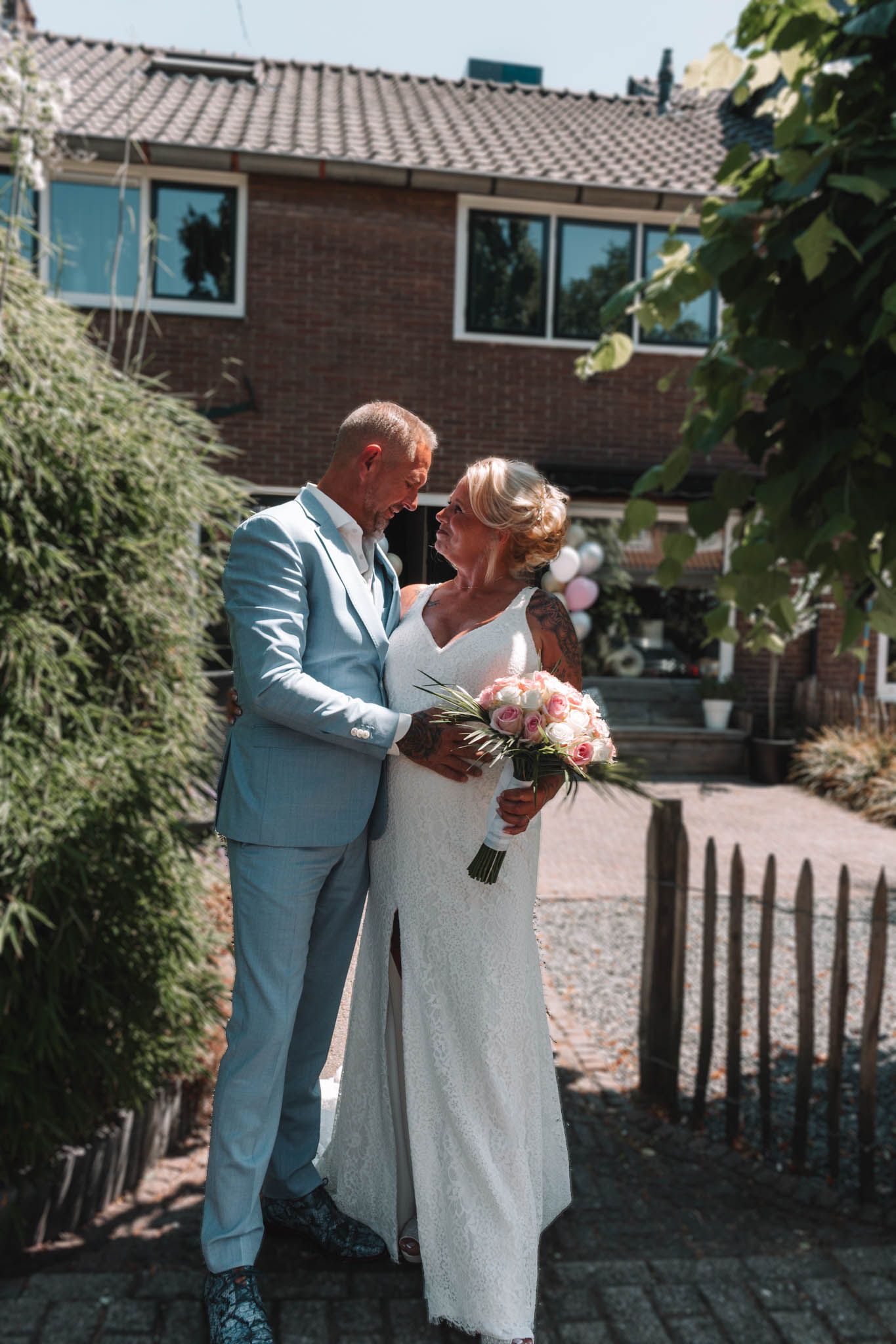 Couple in wedding attire kissing outdoors, holding a bouquet in a garden driveway.