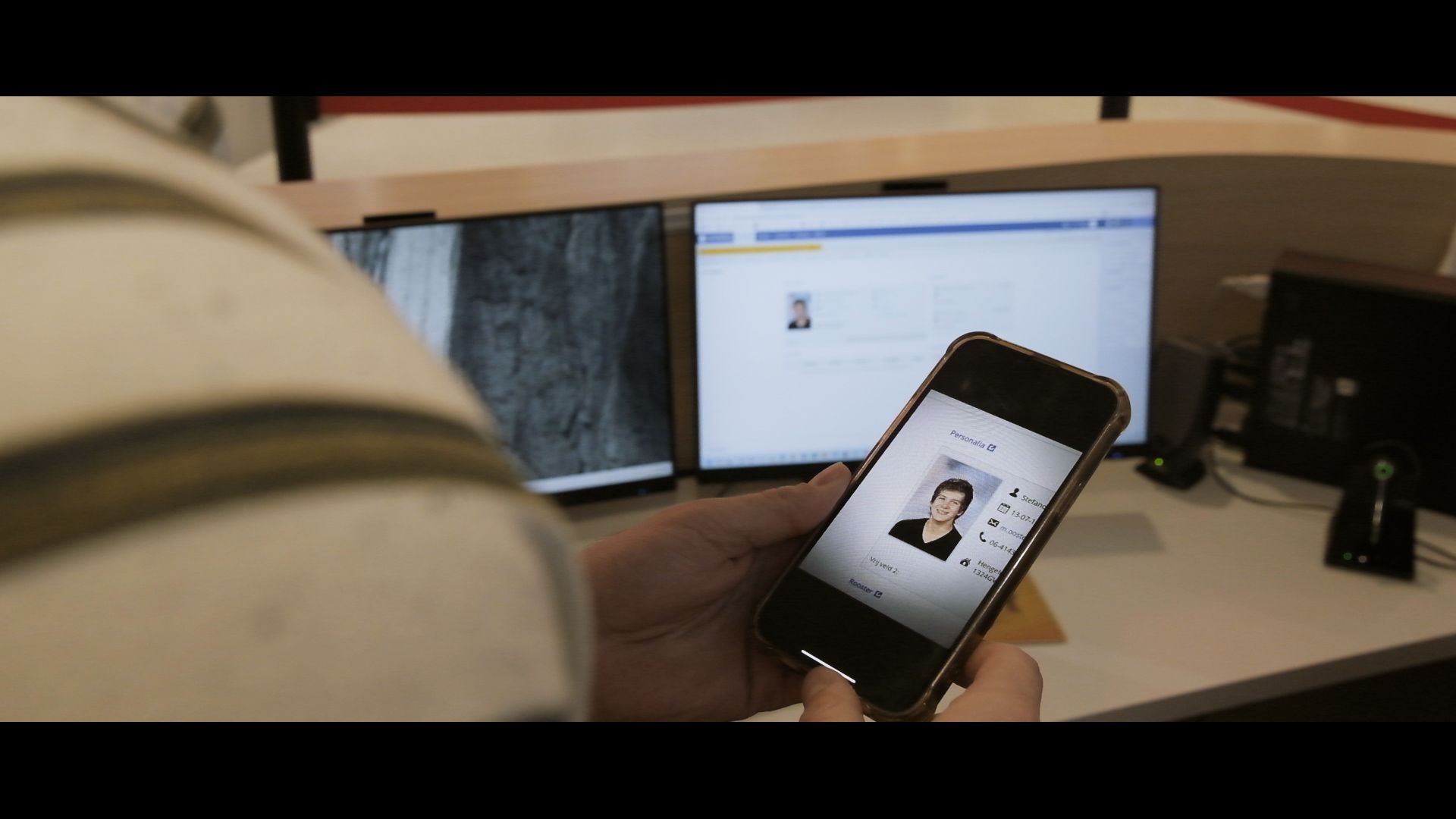 Person holding a smartphone in front of a desk with dual computer monitors.