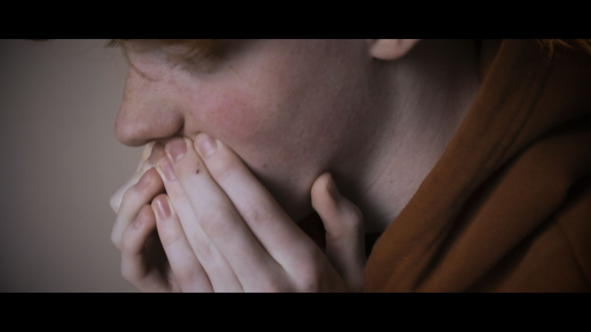 Person in an orange sweater covering their mouth with hands, looking pensive or worried in profile.