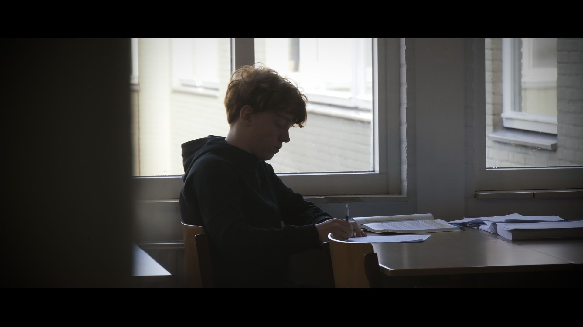 Silhouetted person seated at a desk by a bright window, looking down in a quiet room.