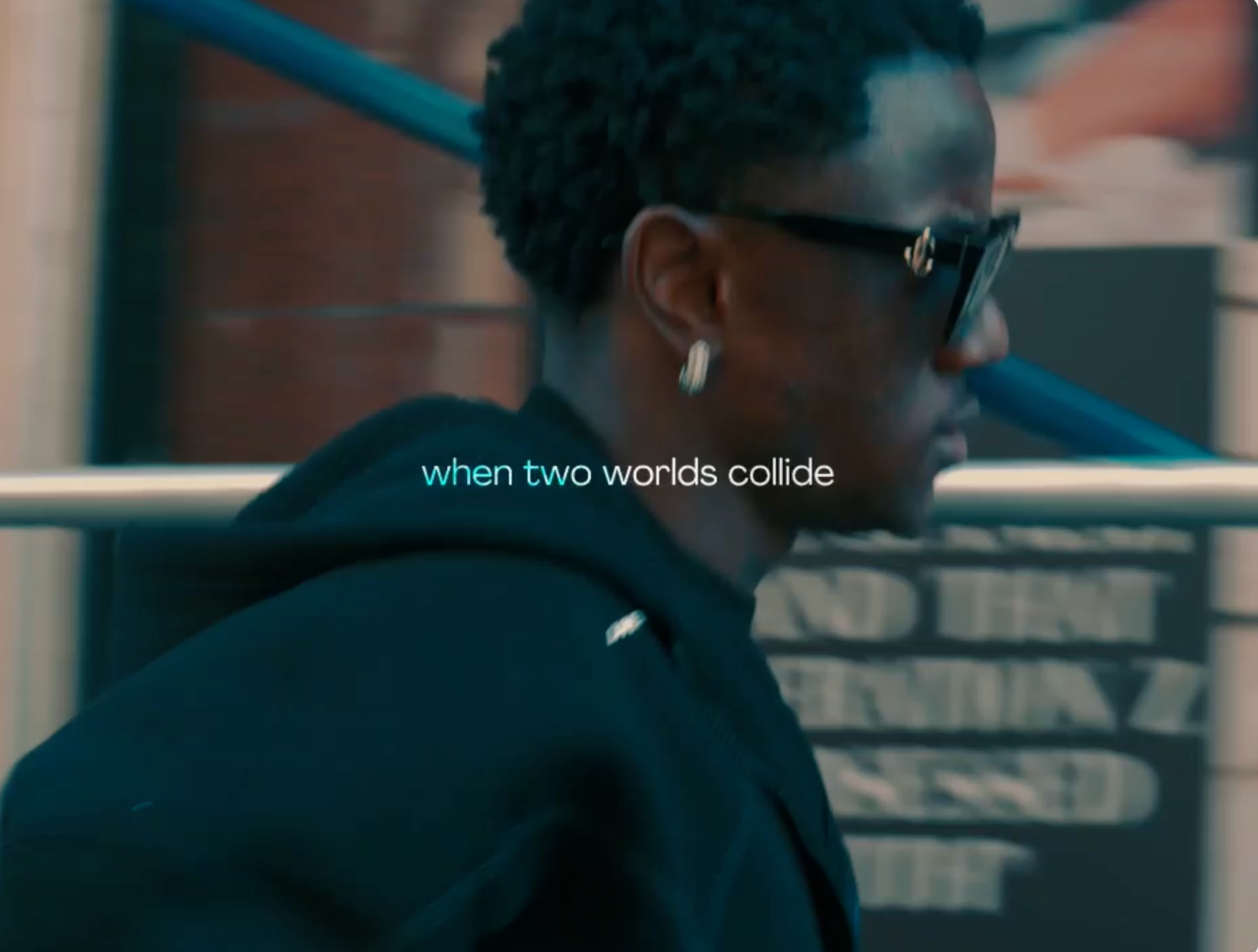 Person in sunglasses and dark hoodie walking past a blurred storefront with text in the background.