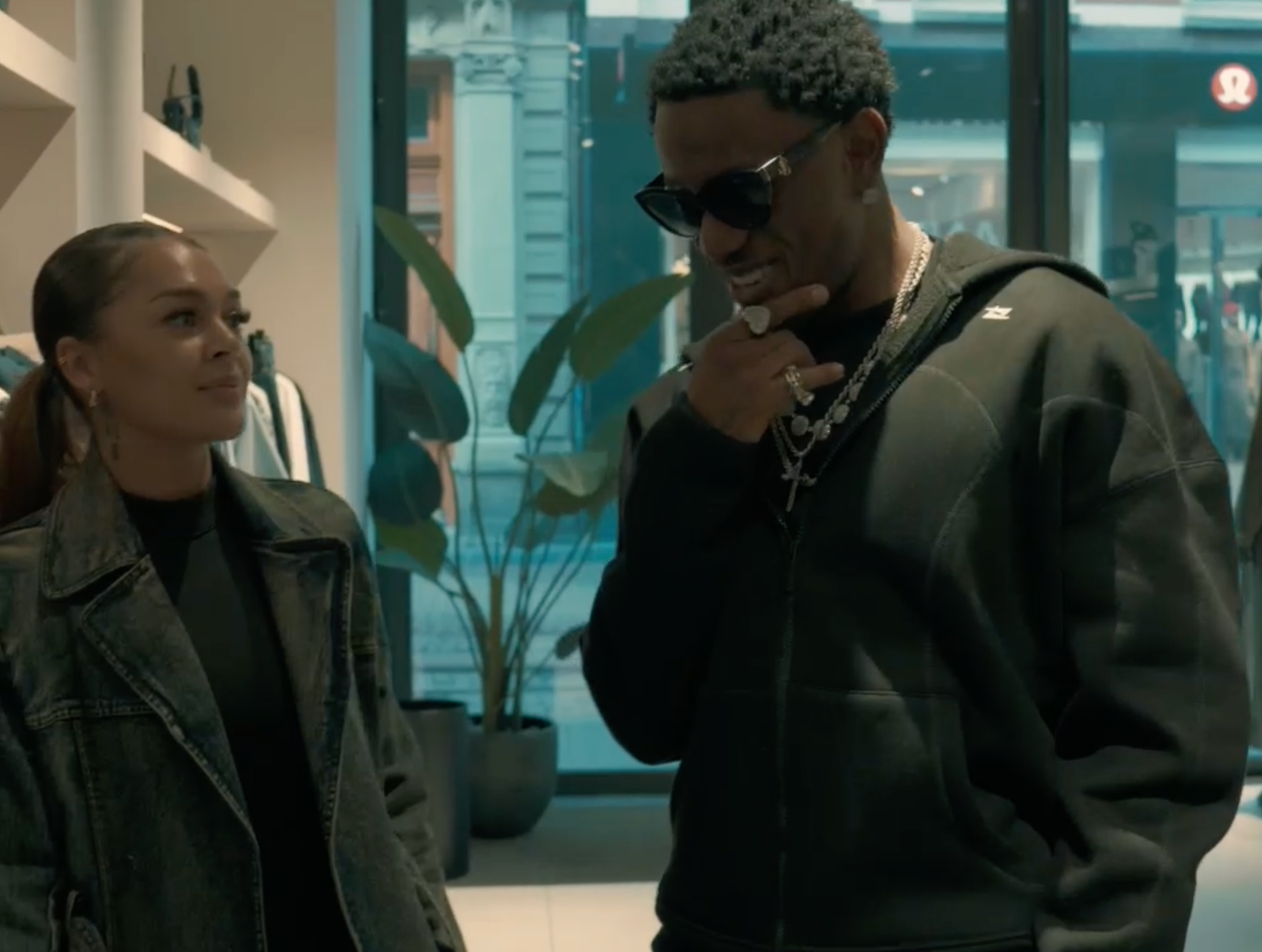 Two people talking in a lobby, one in sunglasses and a dark hoodie, near potted plants and glass doors