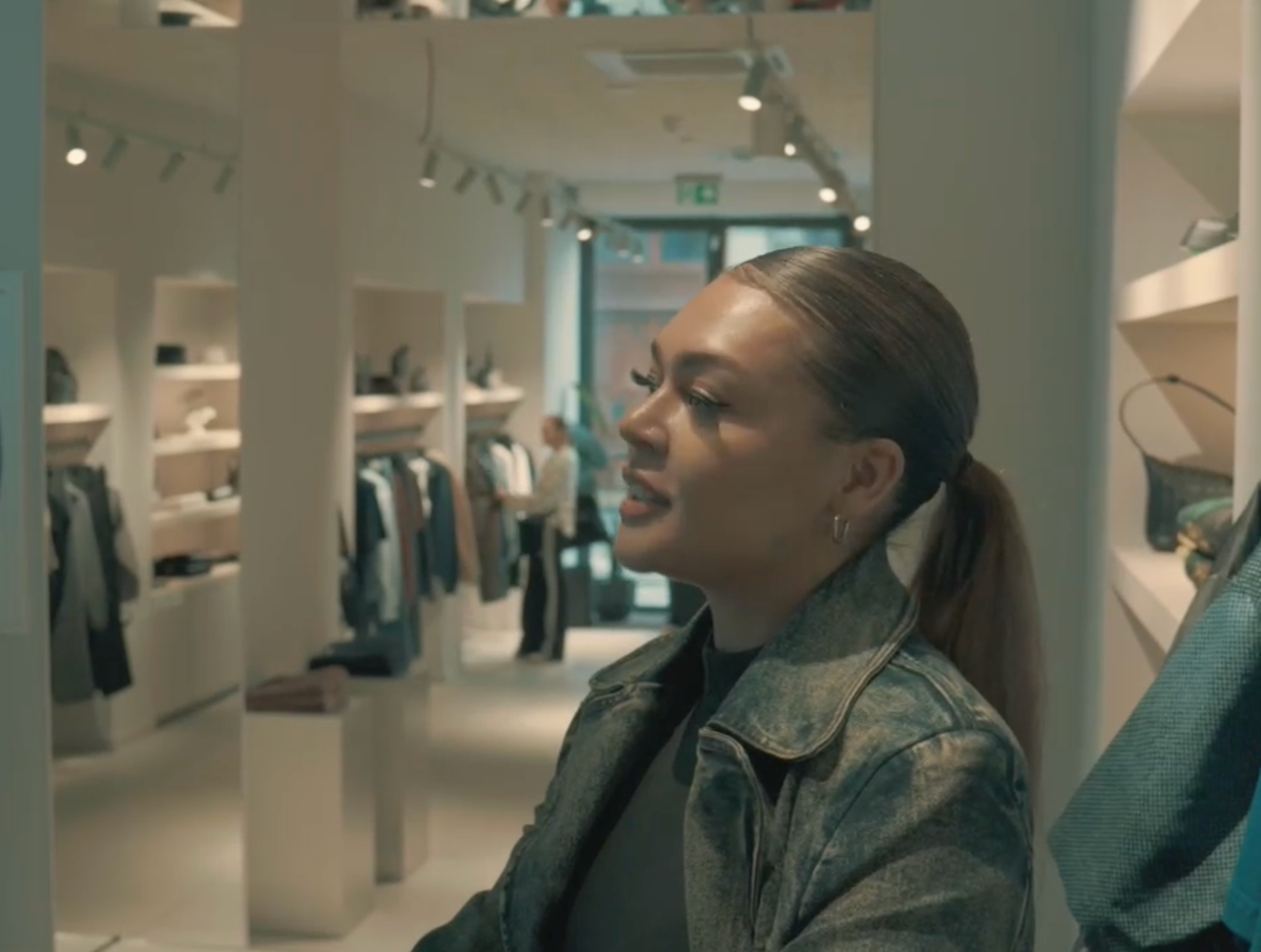 Woman browsing clothing in a modern boutique, wearing a denim jacket and looking to the side.