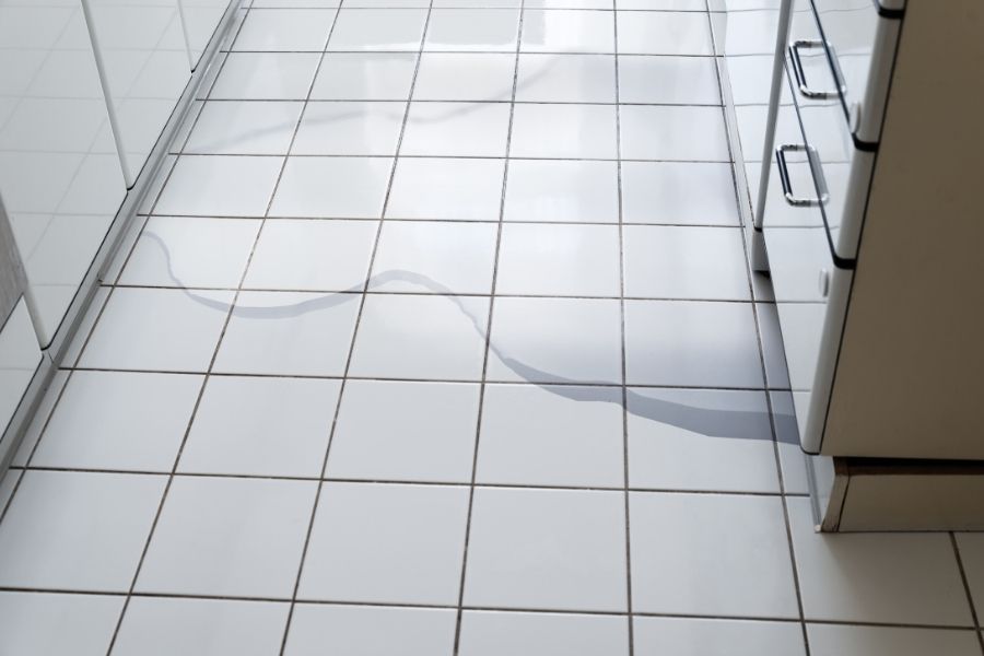 Thin trail of water winding across white kitchen floor tiles from beneath a white appliance