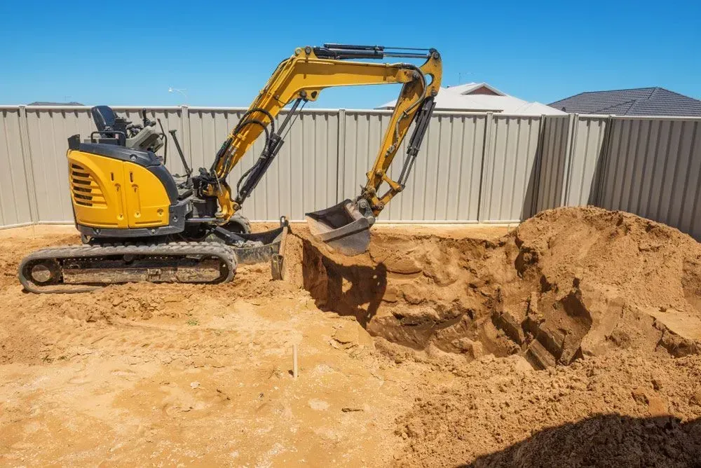 Yellow Excavator Digging in Sand Next to a White Fence Under a Blue Sky — Huntlys Heavy Equipment In Gracemere, QLD