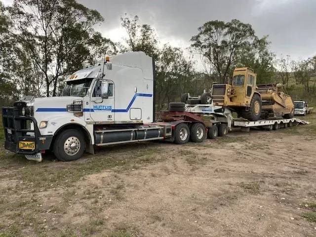 White Truck Pulling a Lowboy Trailer Carrying Yellow Construction Equipment — Huntlys Heavy Equipment In Gracemere, QLD