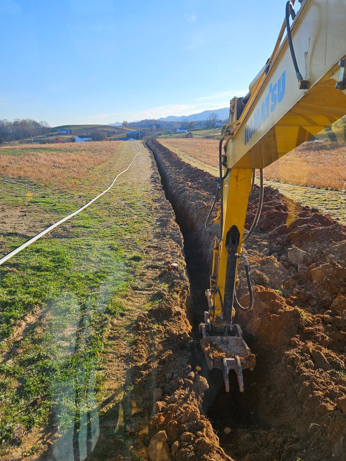 Excavator digging a long trench through a grassy field under a clear blue sky.