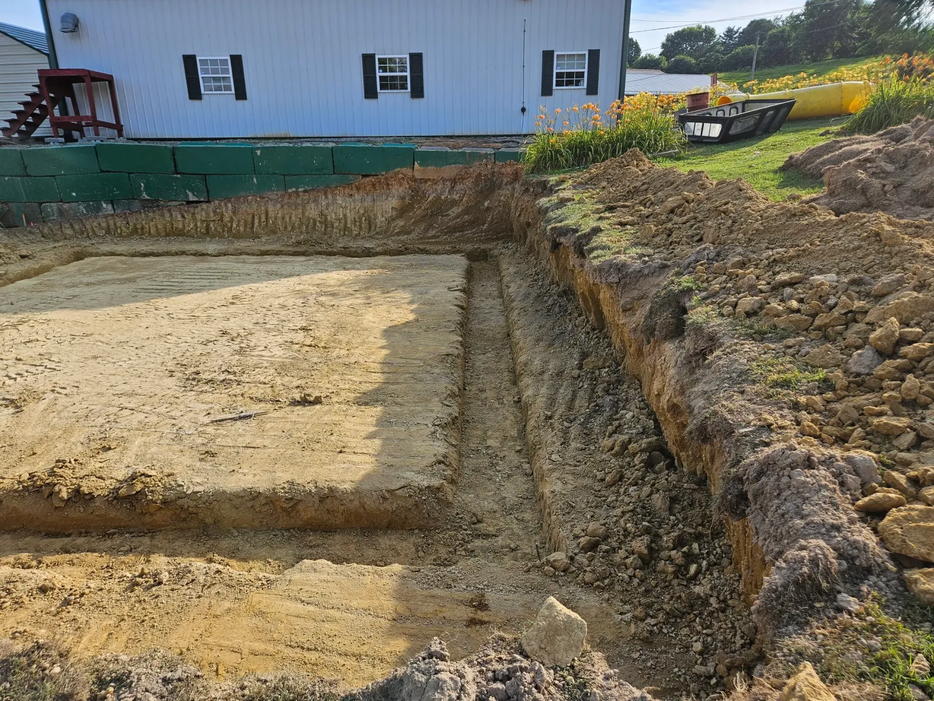 Excavated foundation site beside a white house, with dirt trenches and a sloped soil embankment