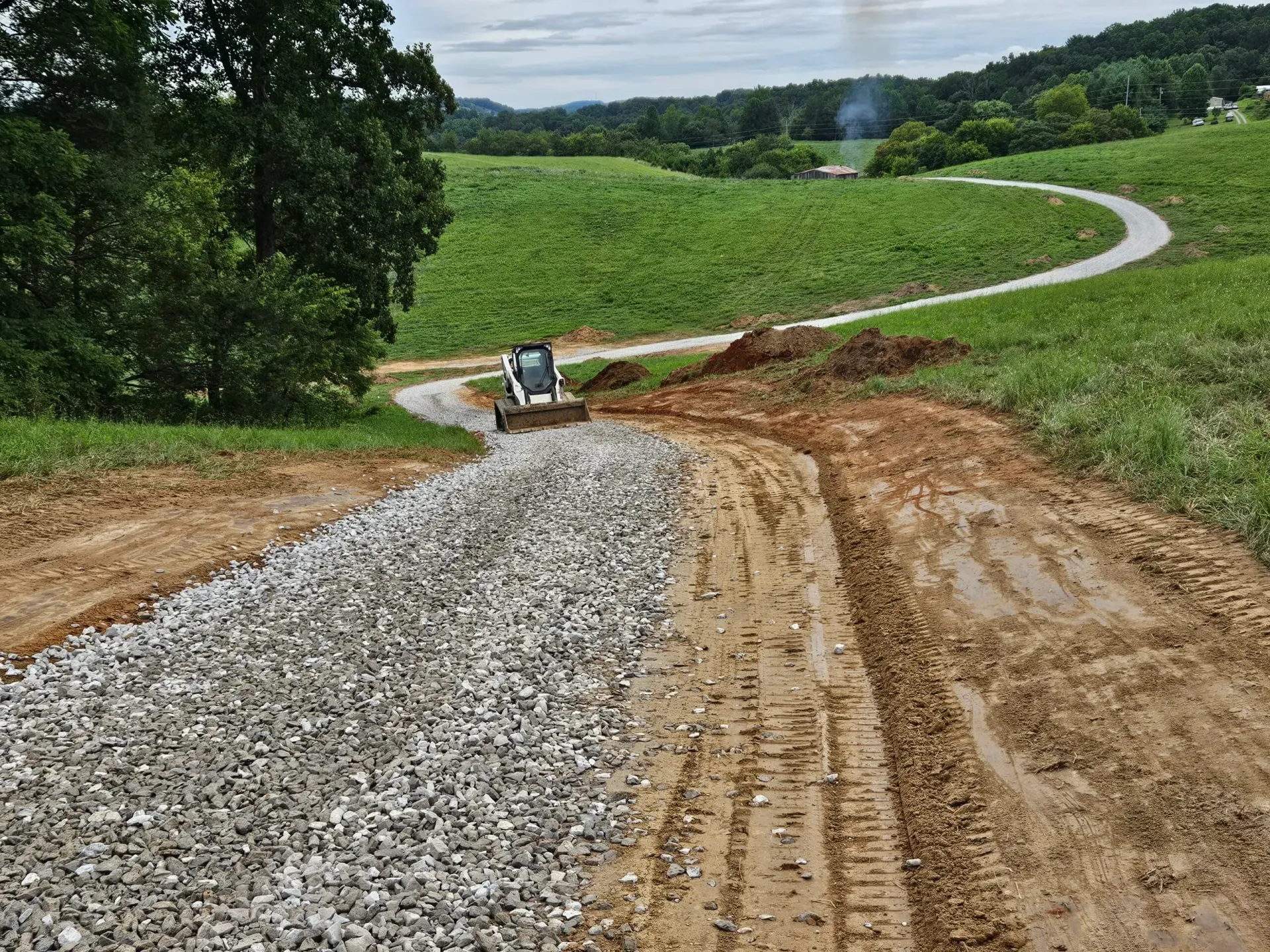 Gravel path under construction winding through green fields, with a small excavator near the bend