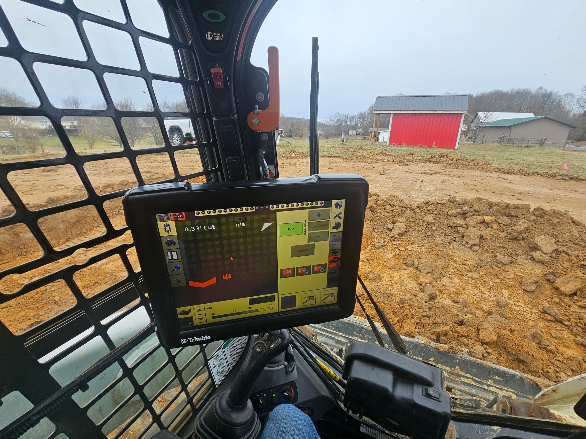 Cab view of a construction machine with a control monitor, overlooking a muddy worksite and red barn.