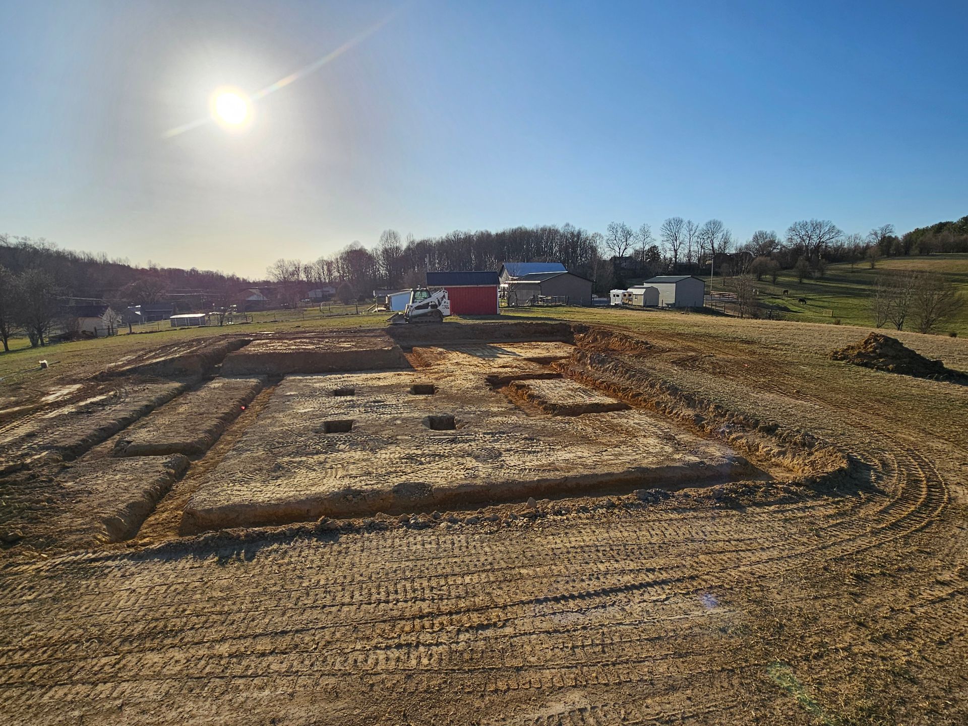 Cleared dirt lot with foundation frames under a bright sun, near trees and parked vehicles