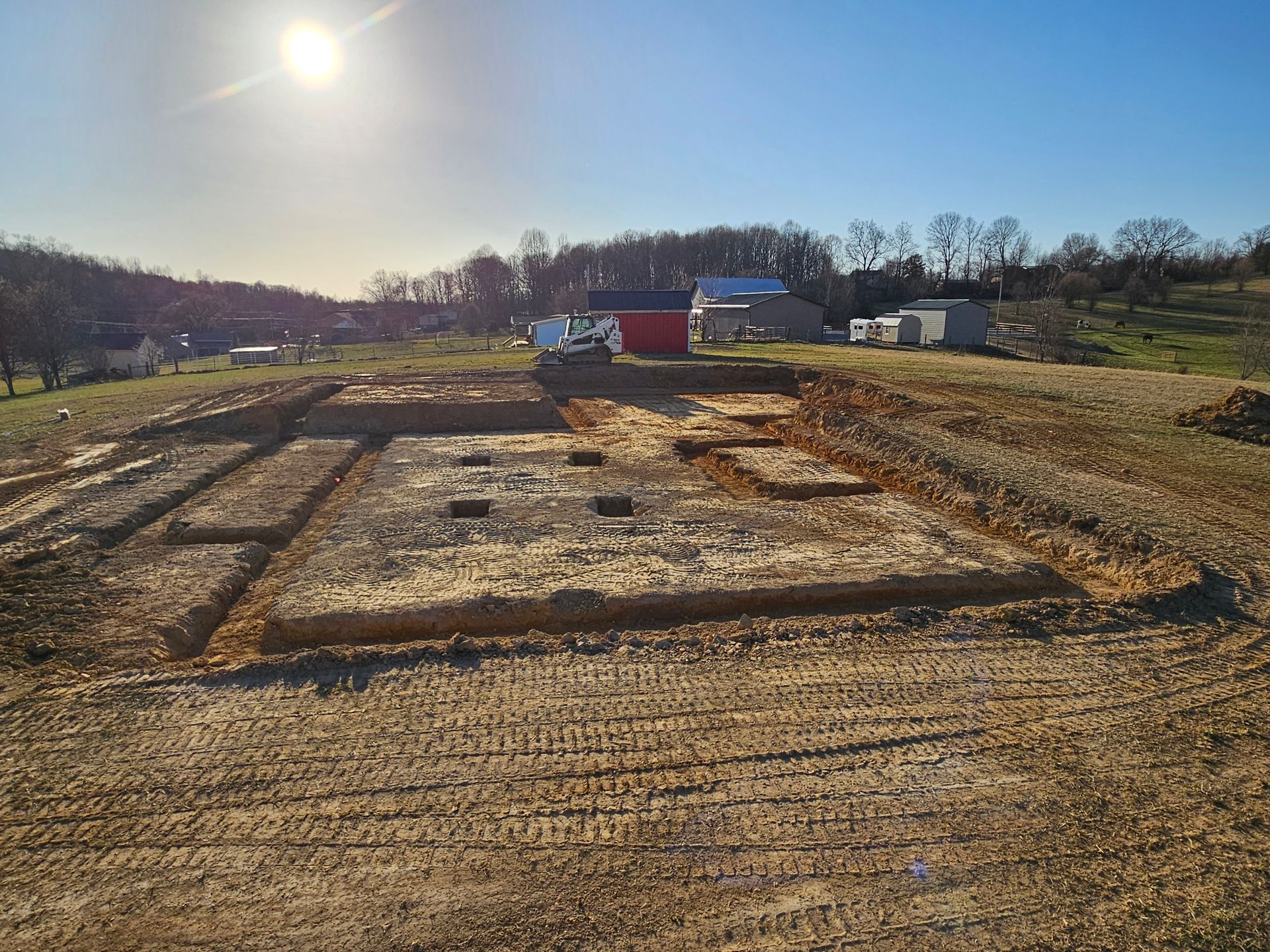 Excavated foundation site in a rural field with dirt trenches, barns, and a bright sun overhead.