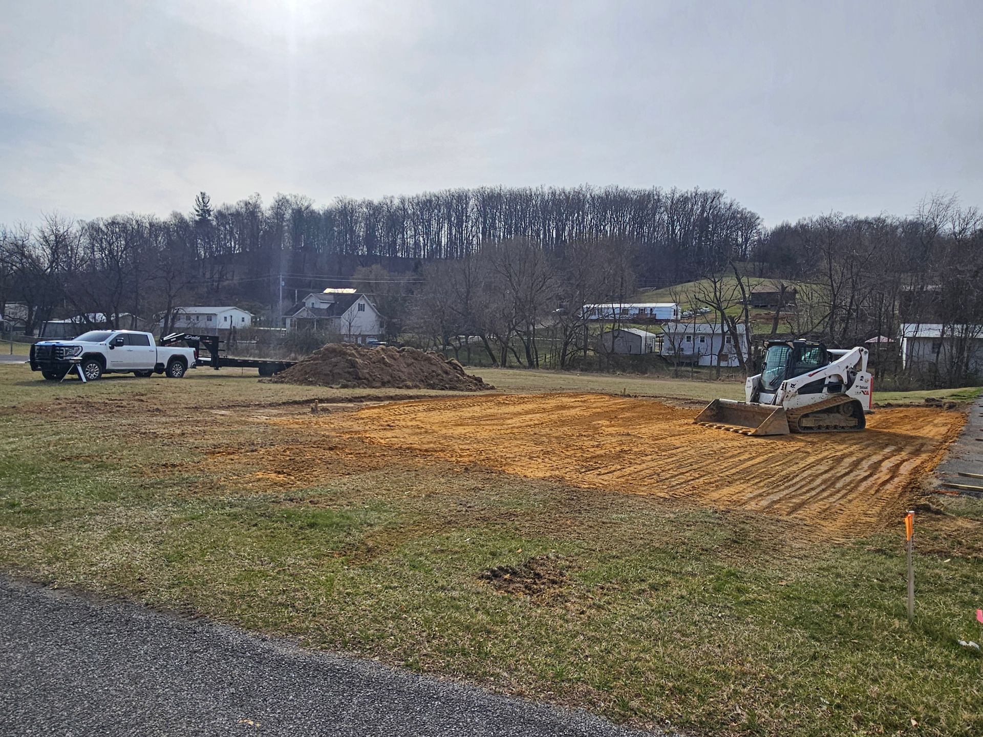 Small excavator grading a dirt lot beside a road, with parked vehicles and trees in the background.