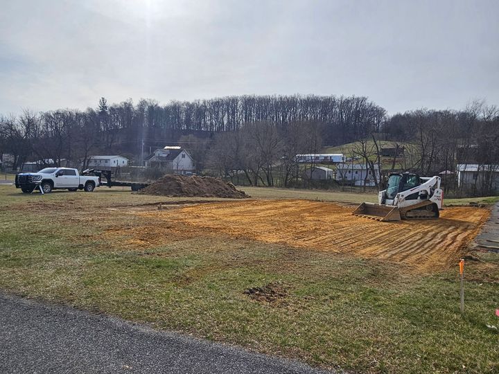 Small excavator grading a dirt lot beside a road, with parked vehicles and trees in the background.