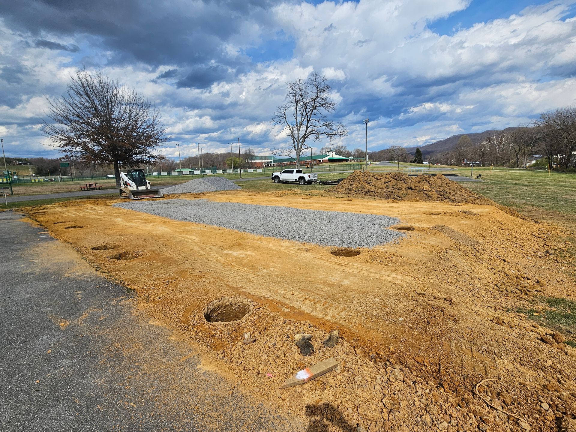 Gravel lot with dirt patches and a few parked vehicles under a cloudy sky near bare trees