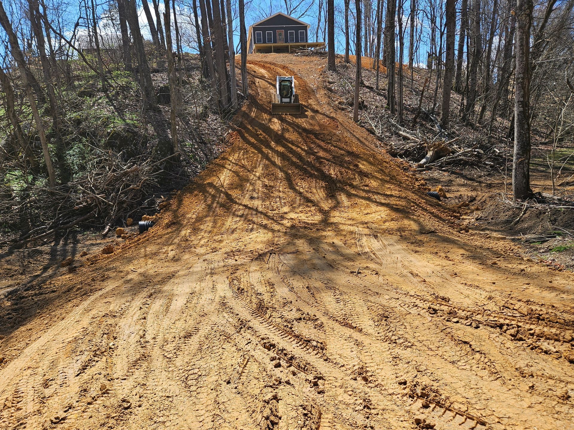 Dirt road climbs a wooded hill to a small house at the top under a clear sky