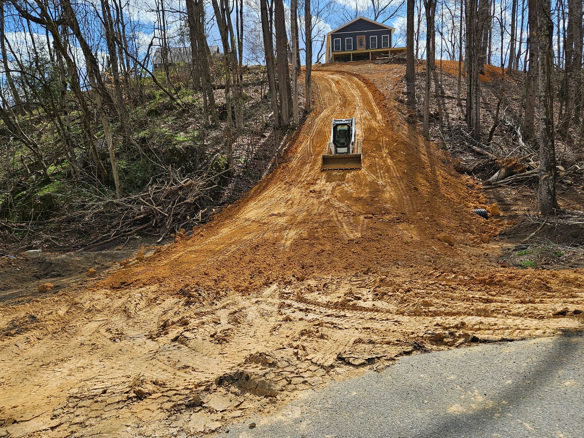 Bulldozer grading a steep dirt hill leading to a small house in a wooded area