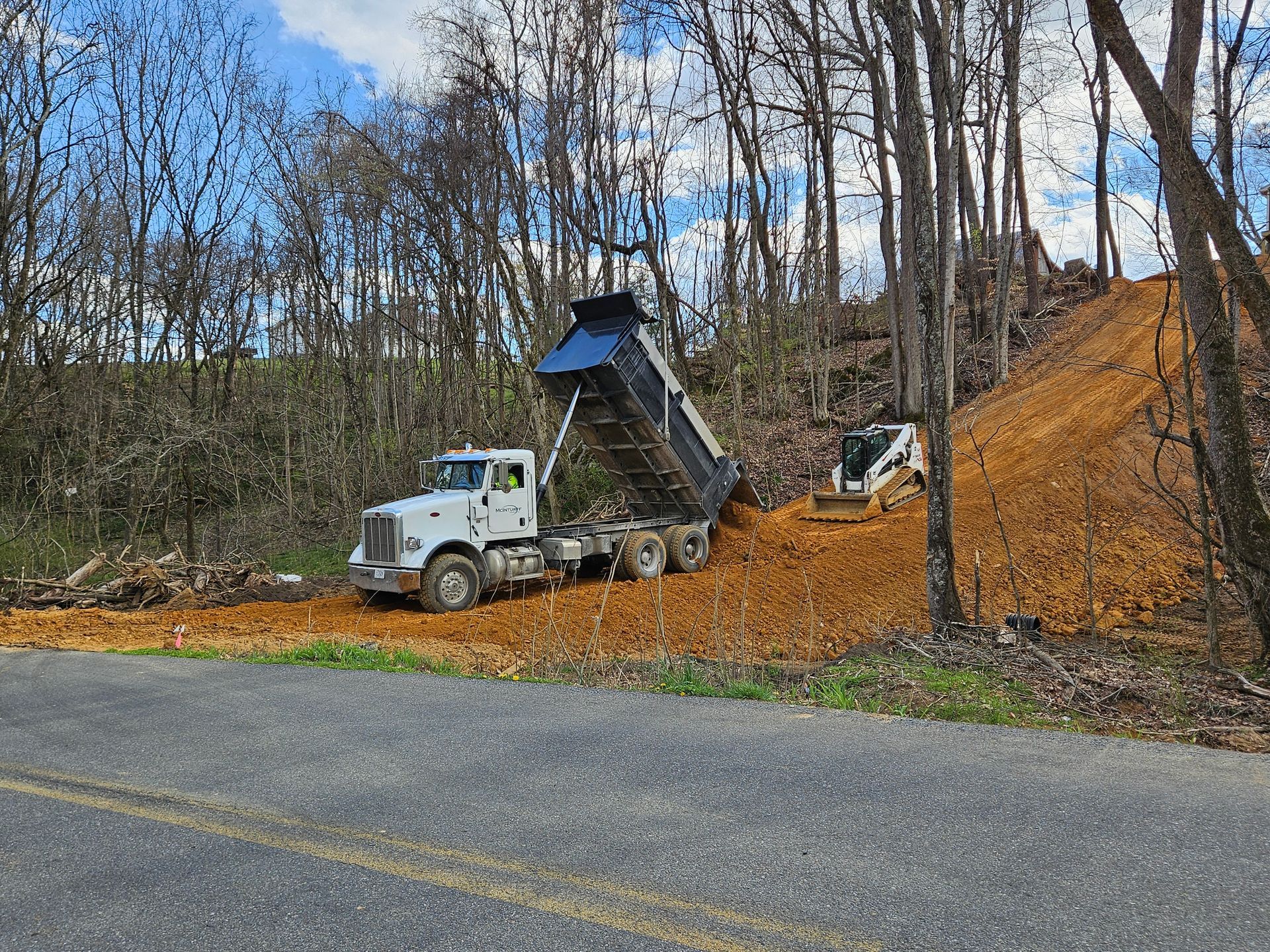 White dump truck unloading mulch on a wooded roadside beside a paved road