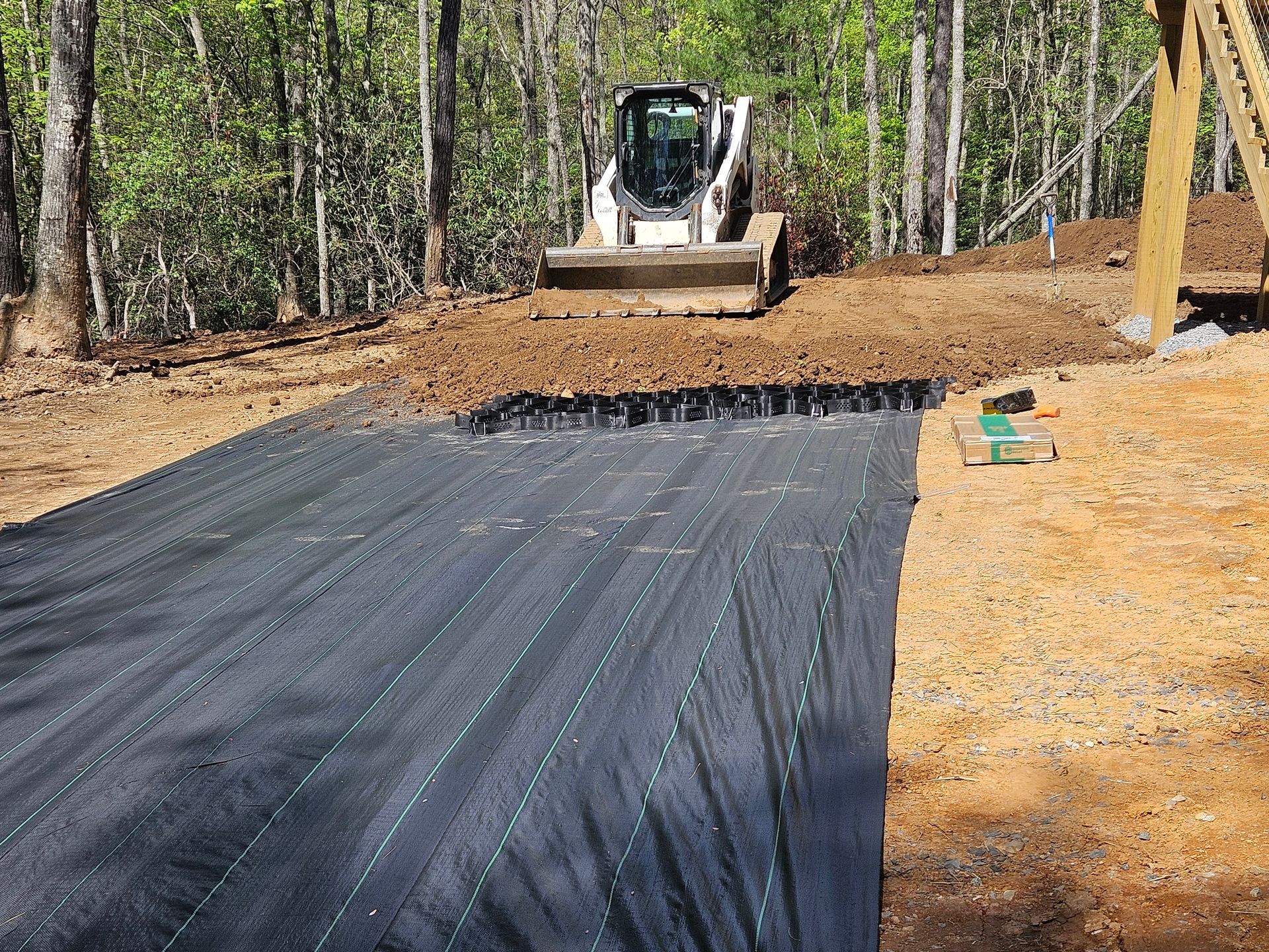 Excavator grading a dirt lot beside a black erosion-control tarp in a wooded area