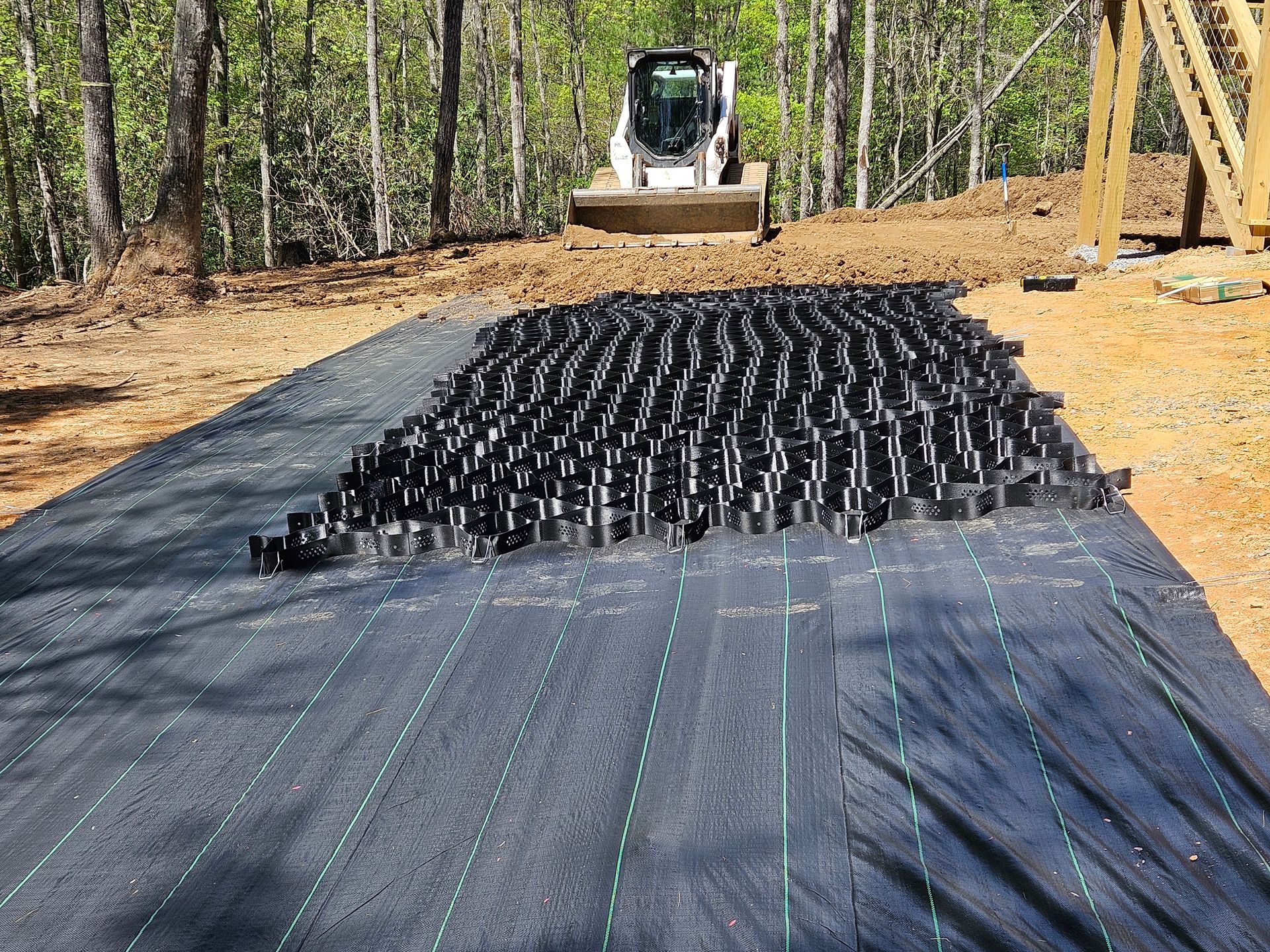 Black fabric mat covered with many small black seedlings at a wooded outdoor worksite with a skid steer in back