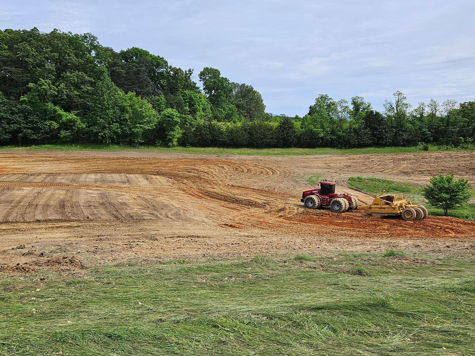 Small tractor plowing a dry field beside a line of green trees under a blue sky.
