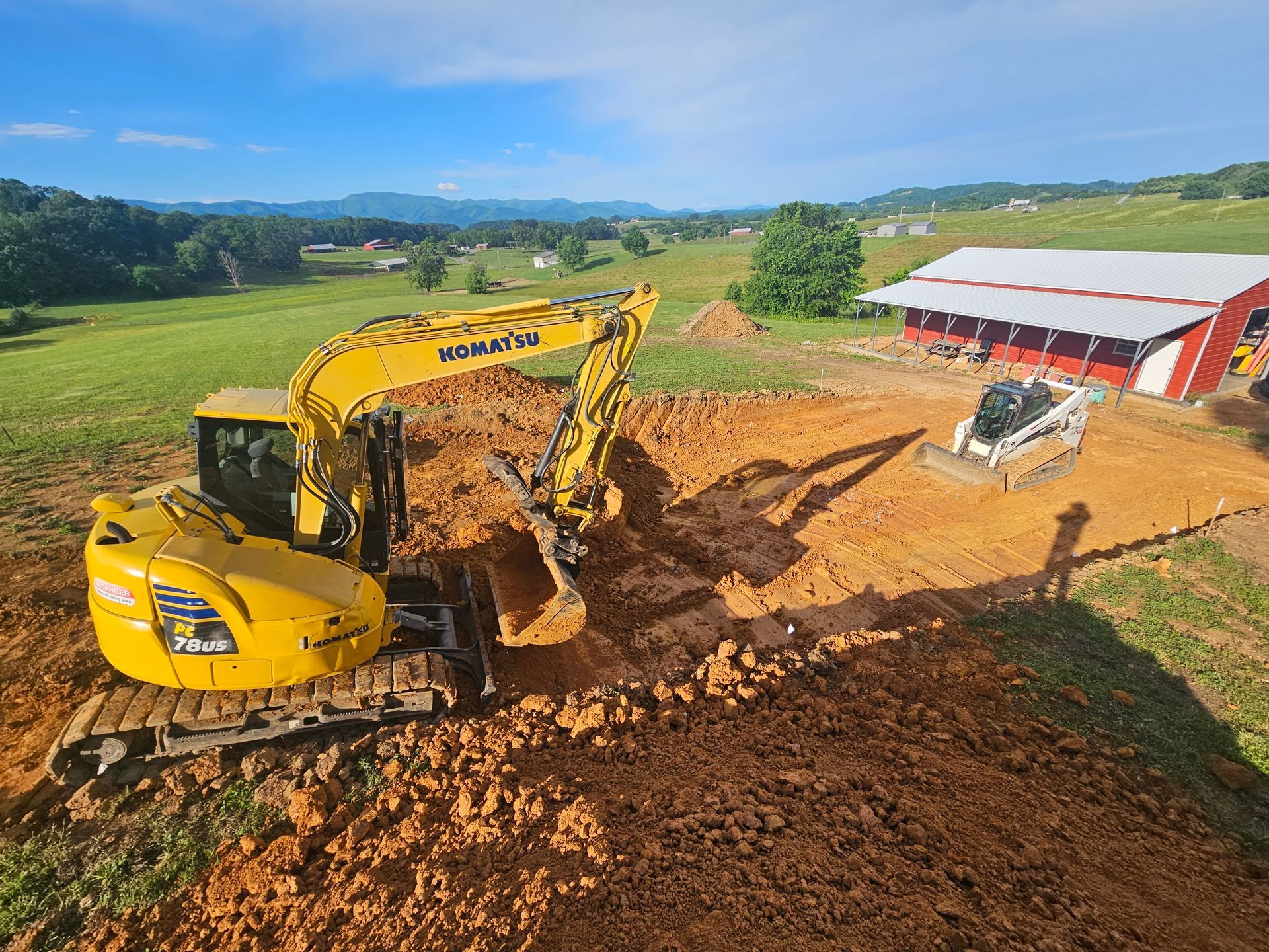Yellow excavator digging red soil beside a white-and-red barn in a rural field.