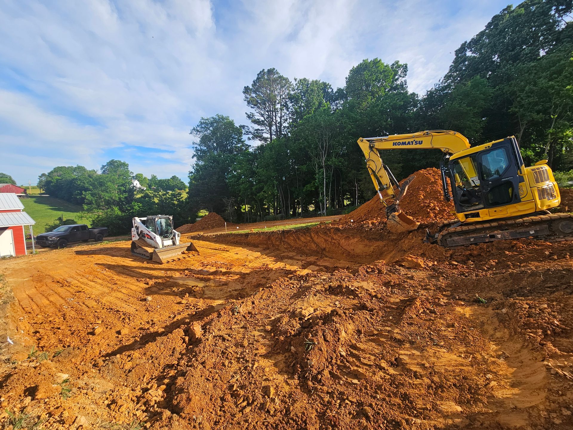 Excavator digging muddy earth on a construction site with a white utility vehicle nearby and trees in the background