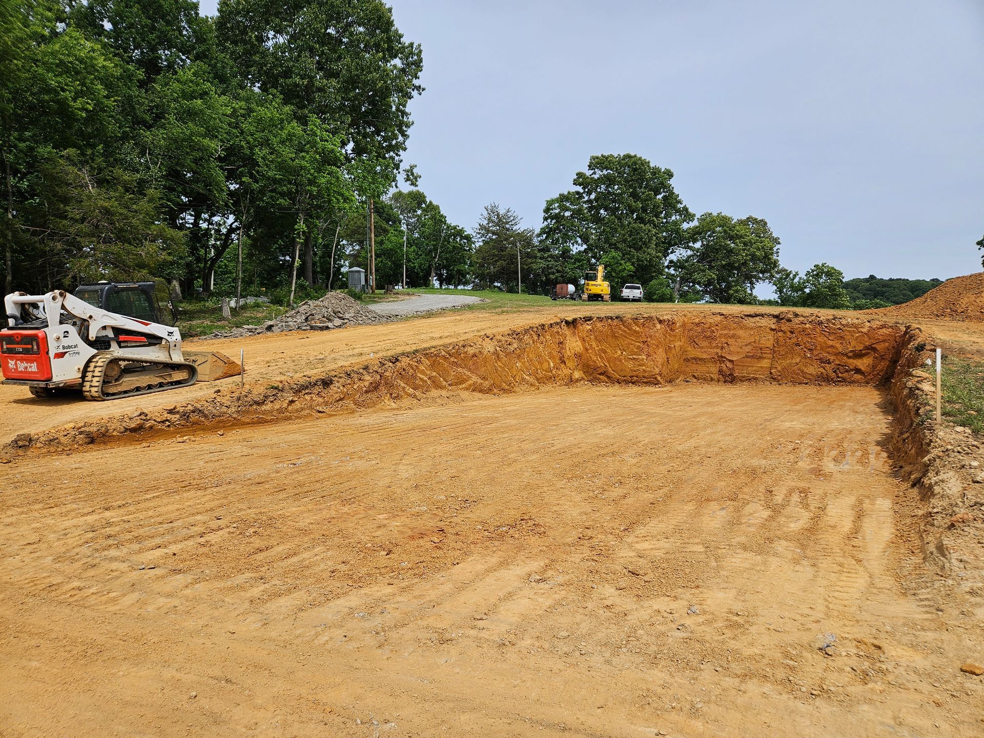 Construction site with excavator grading a large dirt excavation beside trees and a road