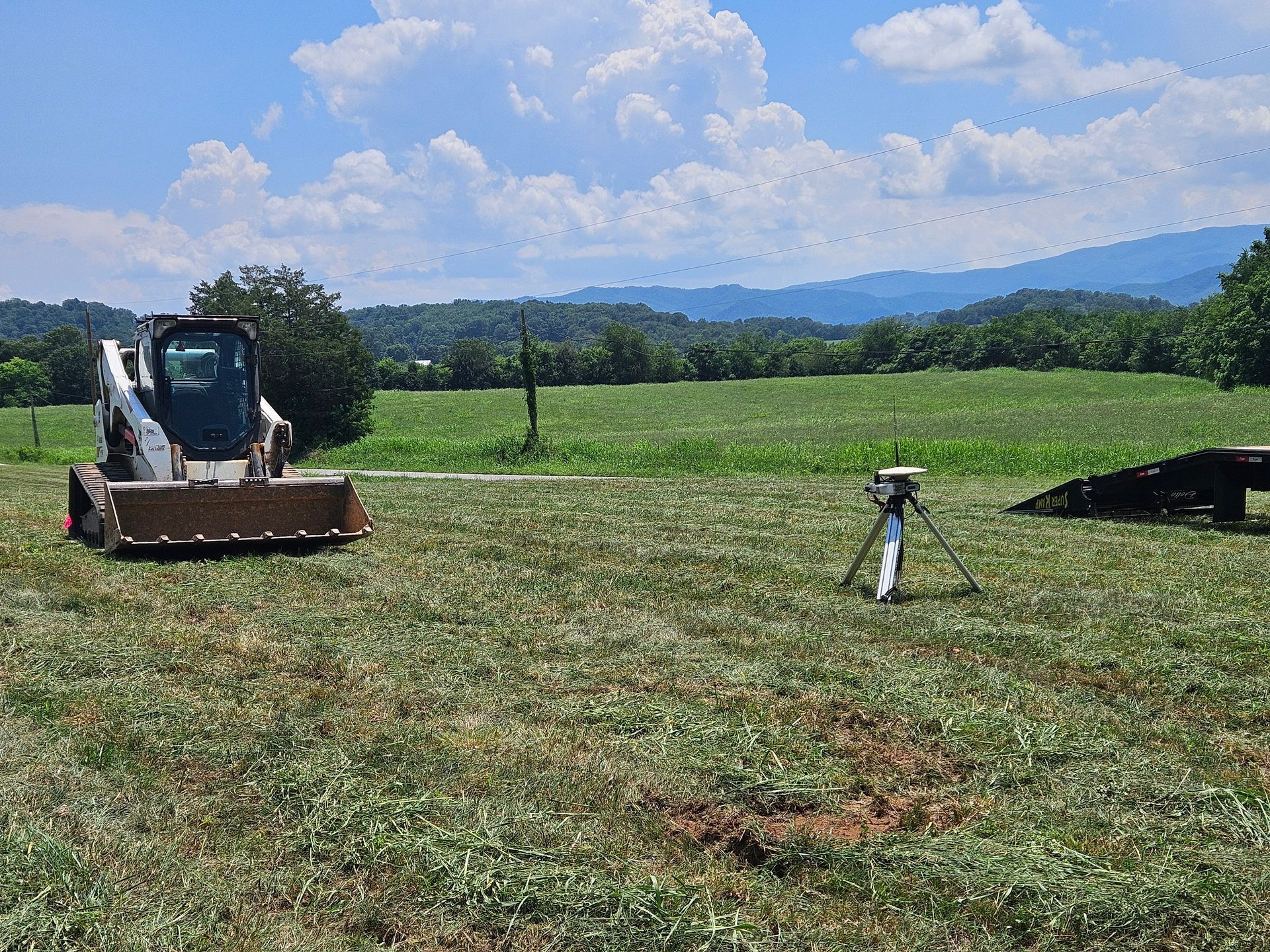 Skid steer on a grassy field with a tripod in the center and mountains under a cloudy blue sky.