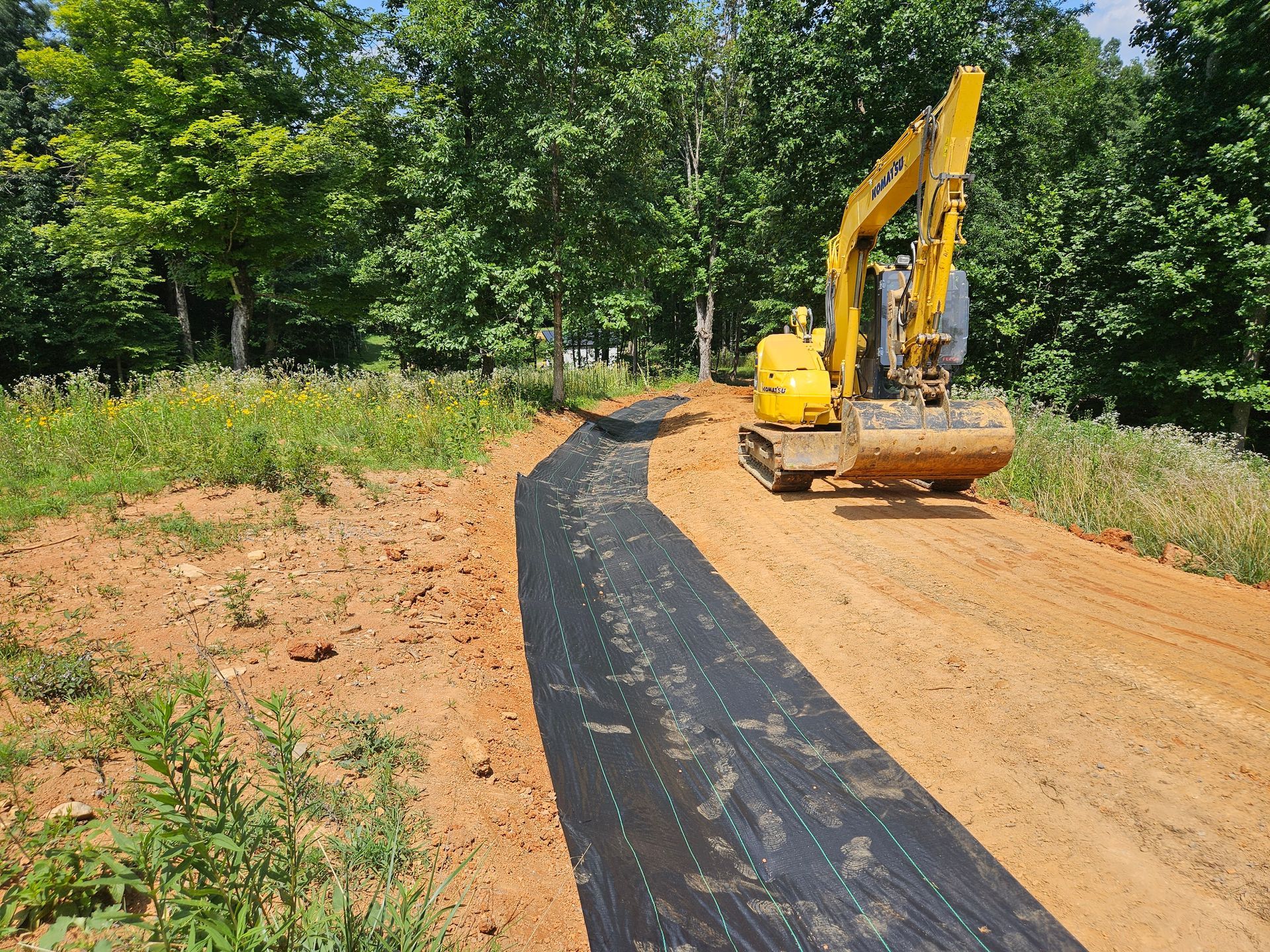 Yellow excavator paving a black road through a dirt path beside green trees and grass