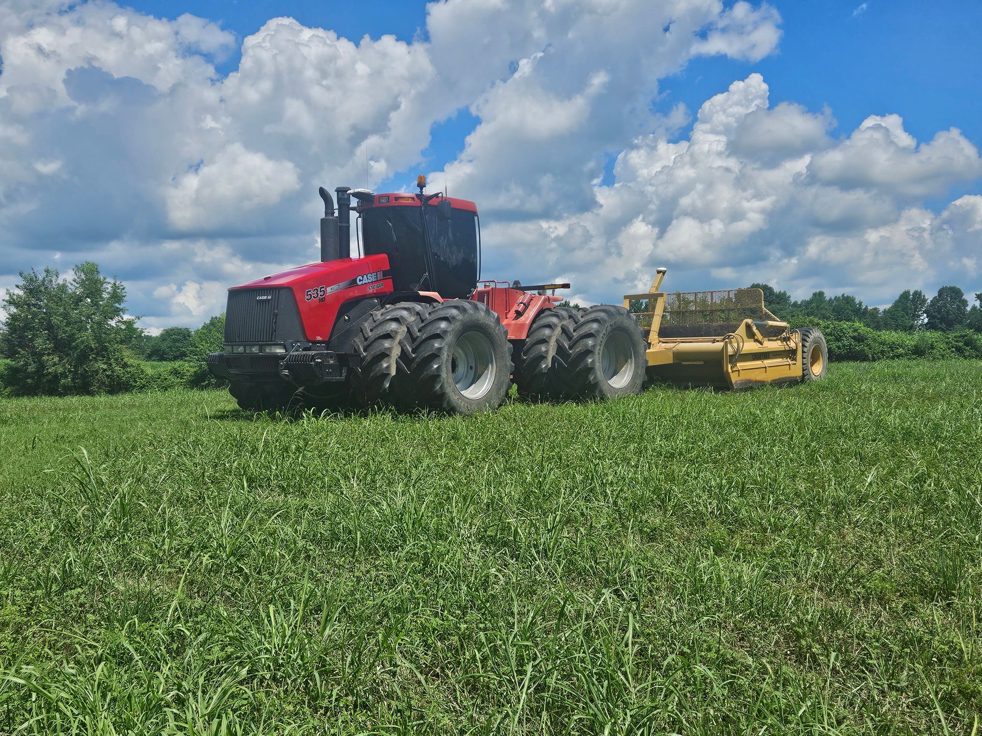 Red tractor towing a yellow farm implement across a green field under a cloudy sky