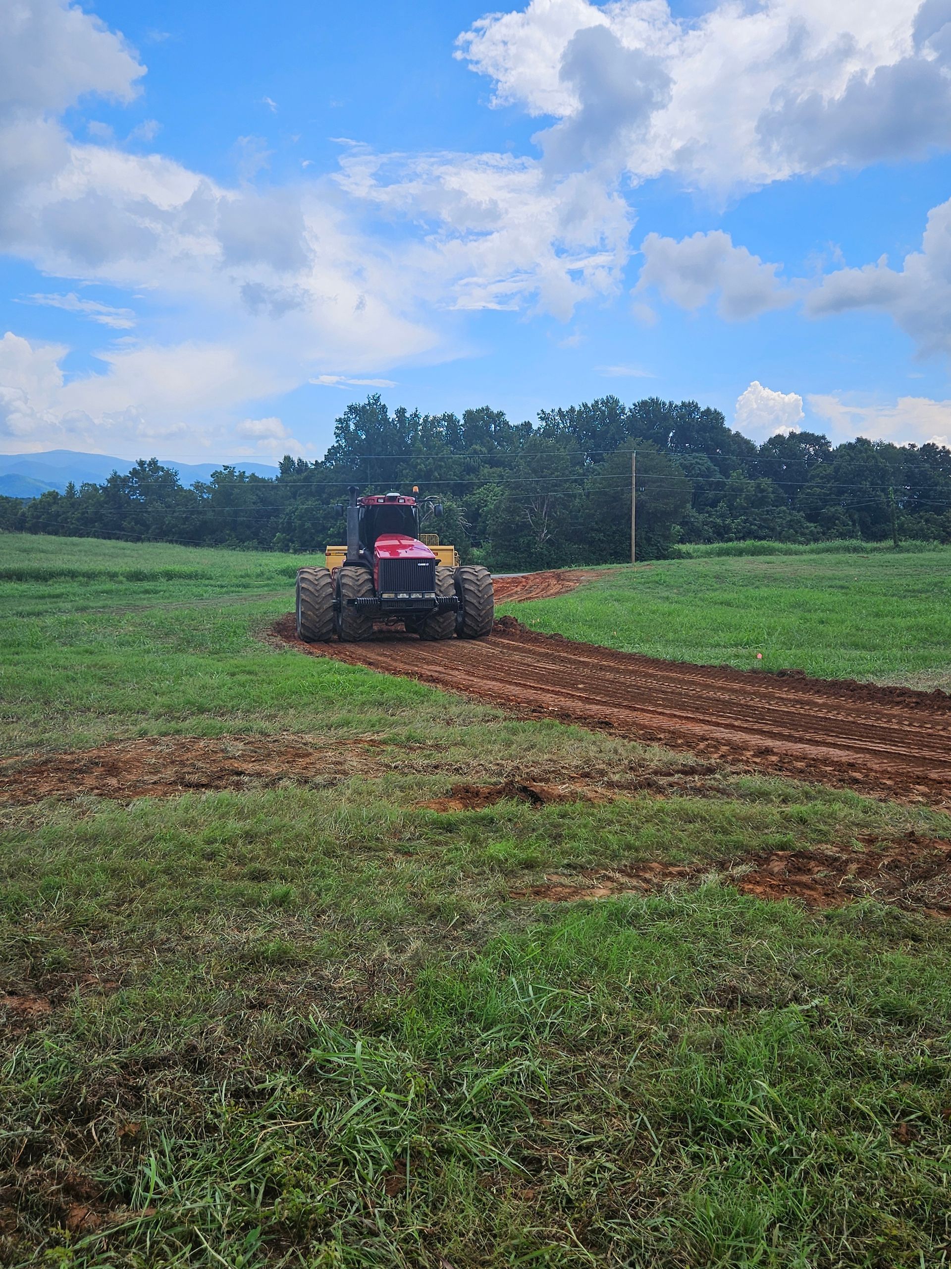 Tractor plowing a muddy field under a cloudy blue sky, with green crops in the background