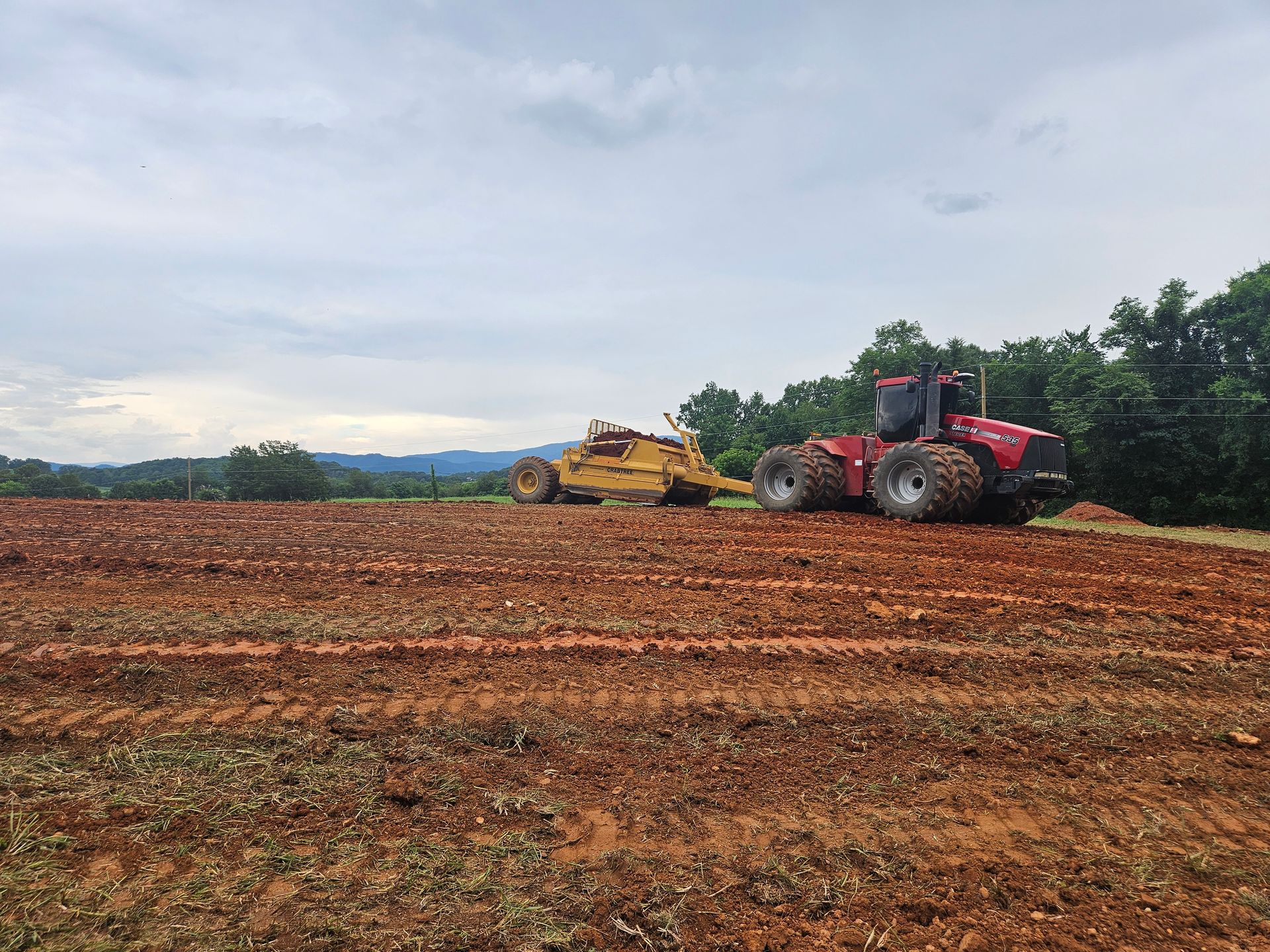 Red tractor and yellow harvester working in a muddy field under a cloudy sky.