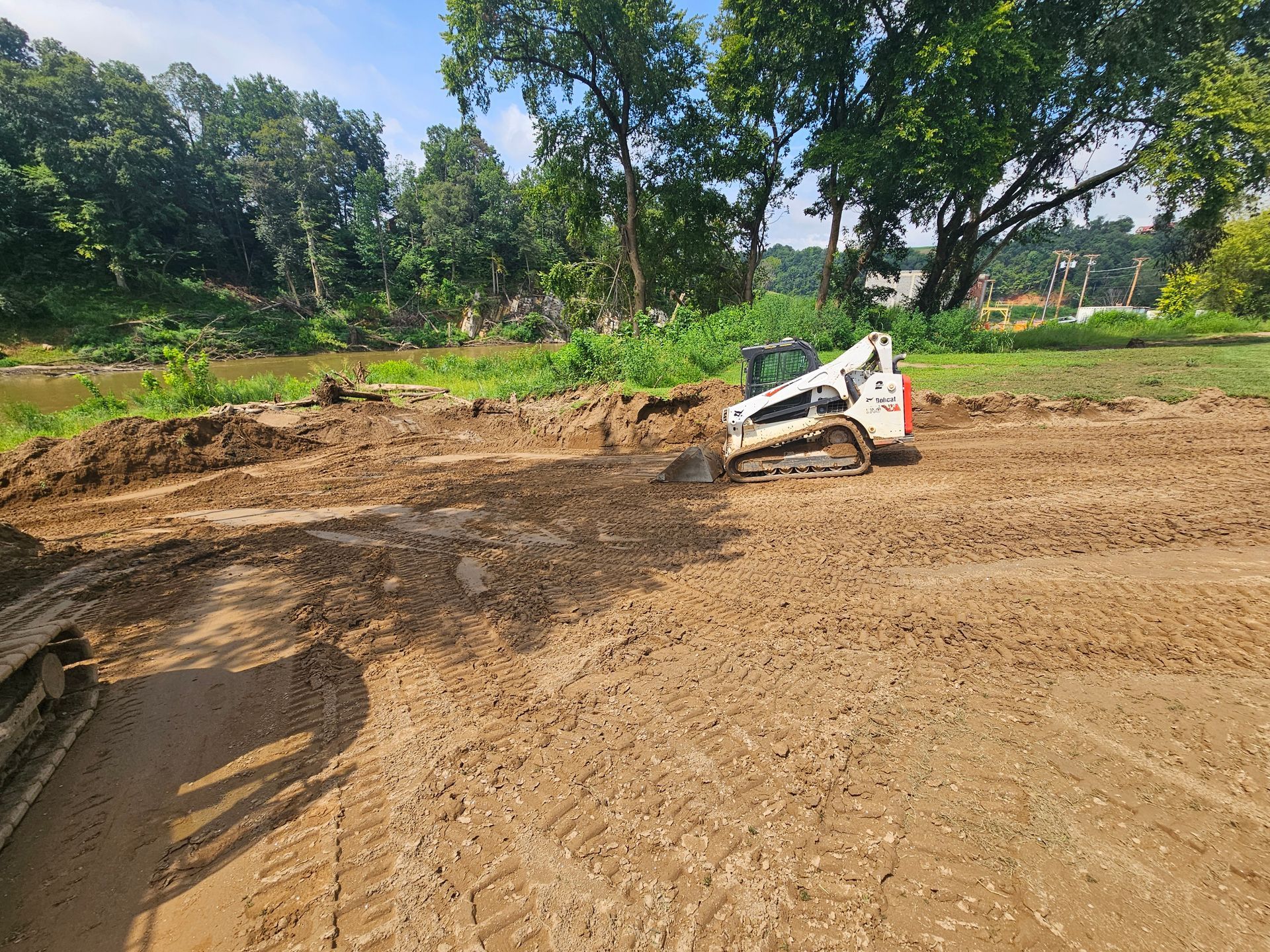 White skid-steer loader grading a dirt site under trees on a sunny day