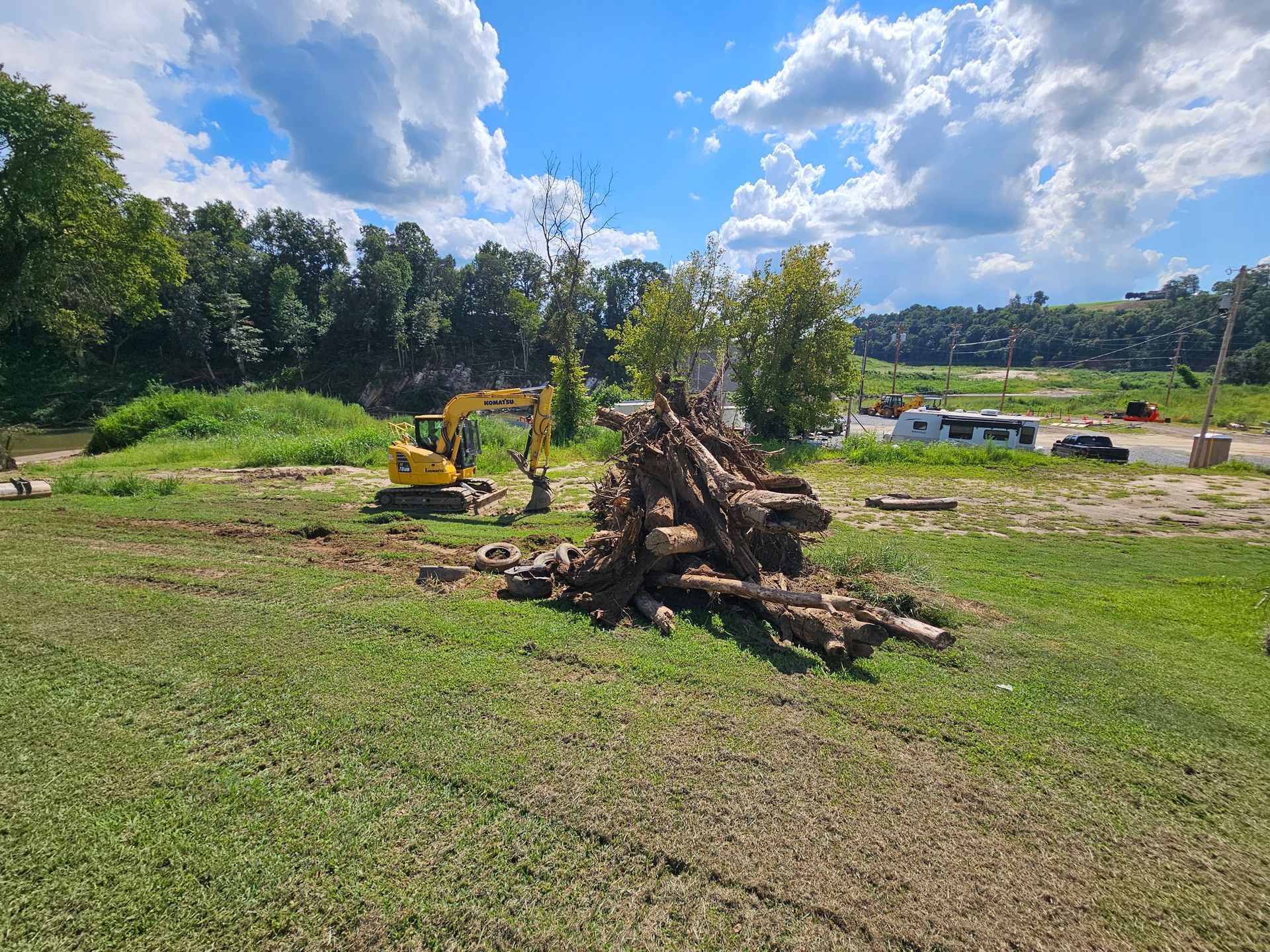 Large woodpile in a grassy field with a yellow excavator and trees under a bright blue sky