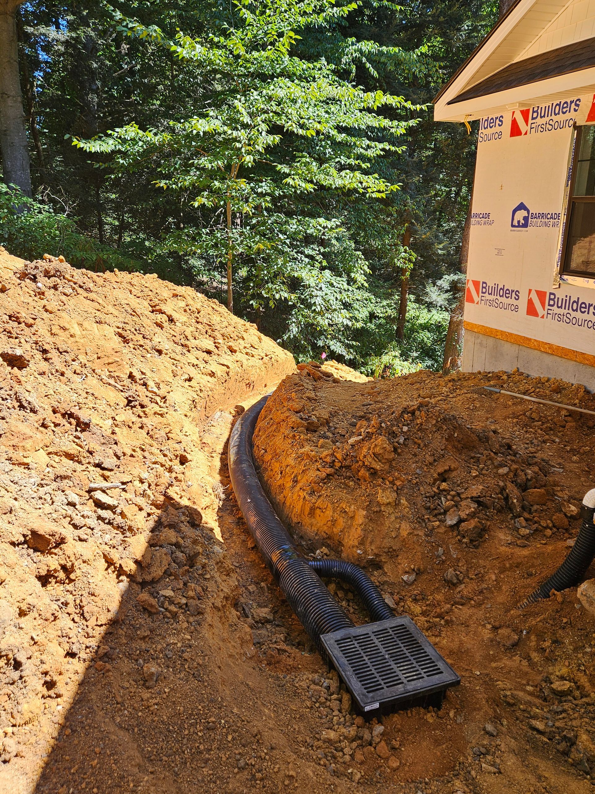 Excavation site with muddy trench, drainage pipe, and construction materials beside a wooded area