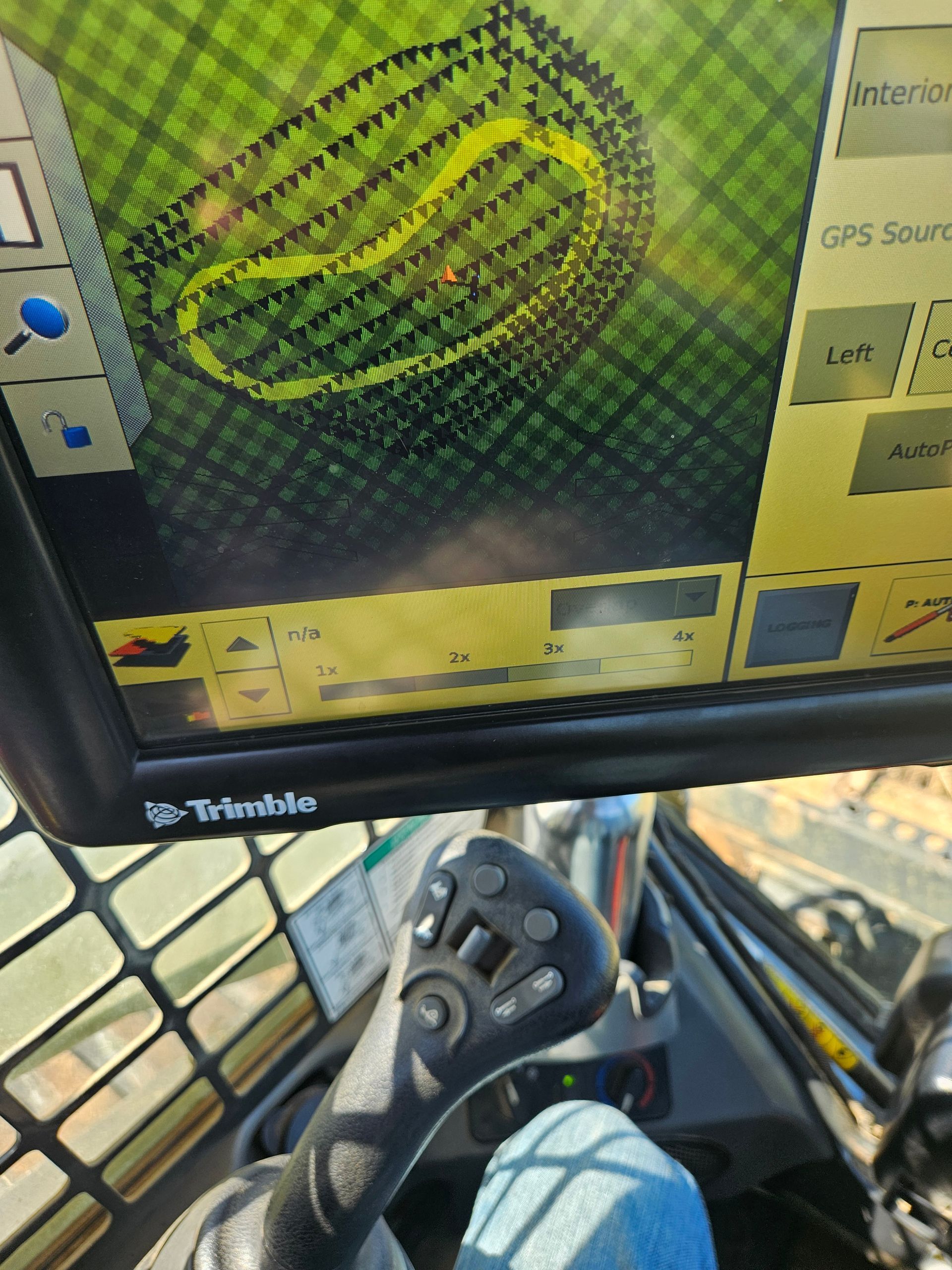 Cab cab view of a Trimble GPS monitor showing a green/yellow guidance map beside a joystick control.