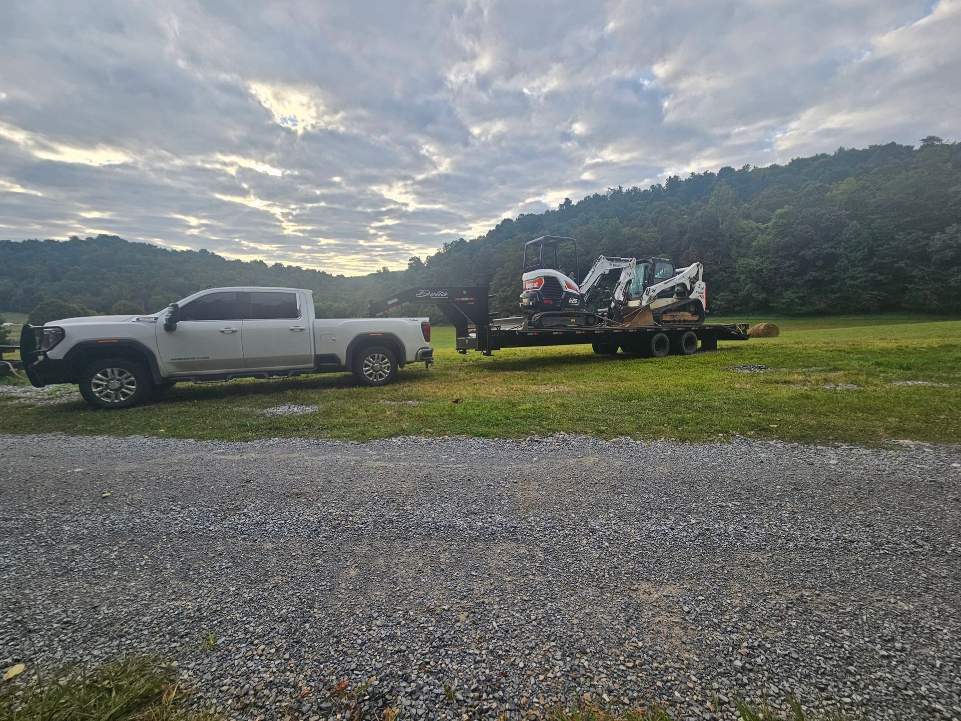 White pickup truck towing an equipment trailer on a gravel lot beside grassy hills under a cloudy sky