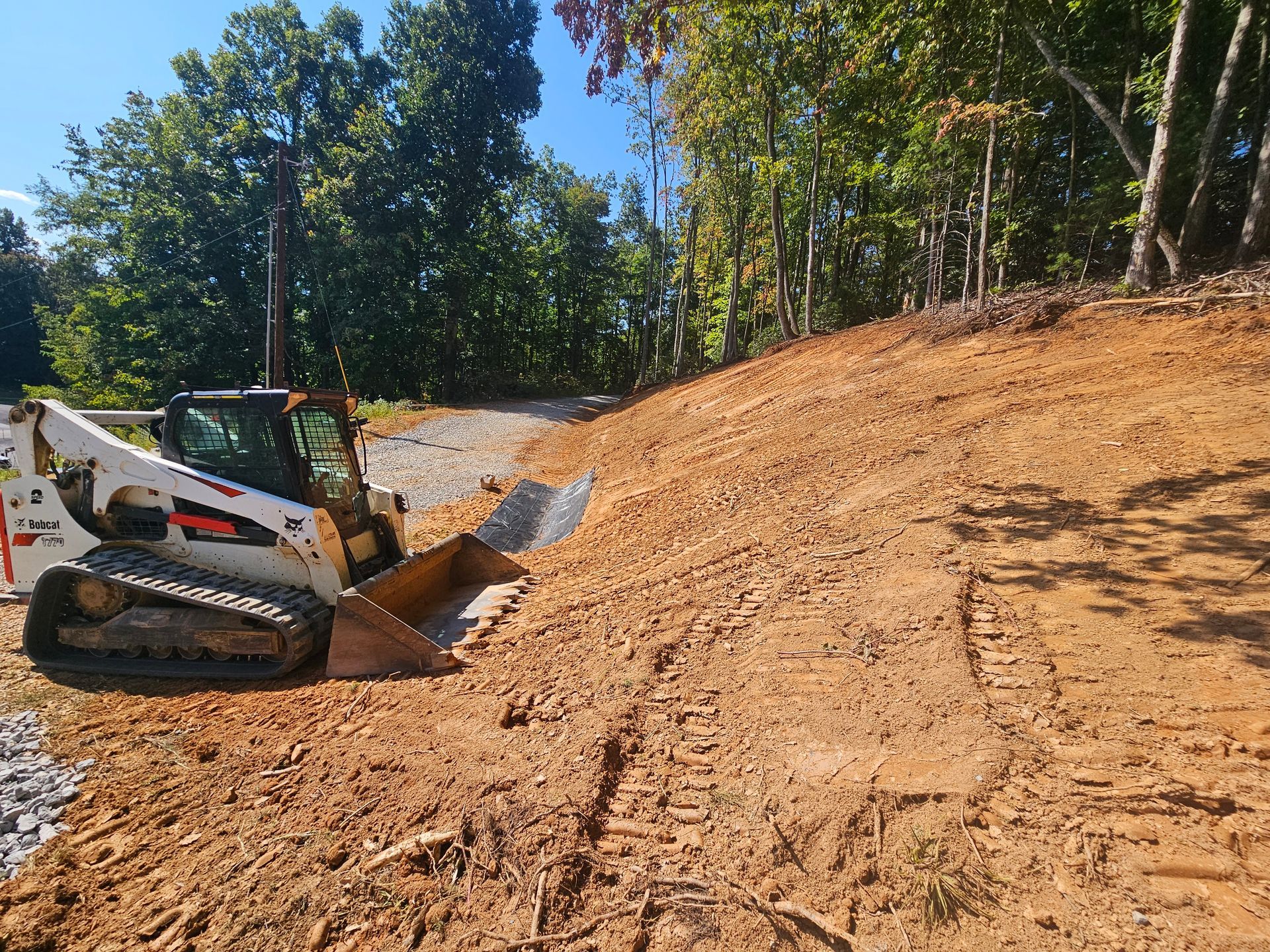 White skid-steer loader leveling dirt on a sloped construction site beside trees and a gravel road