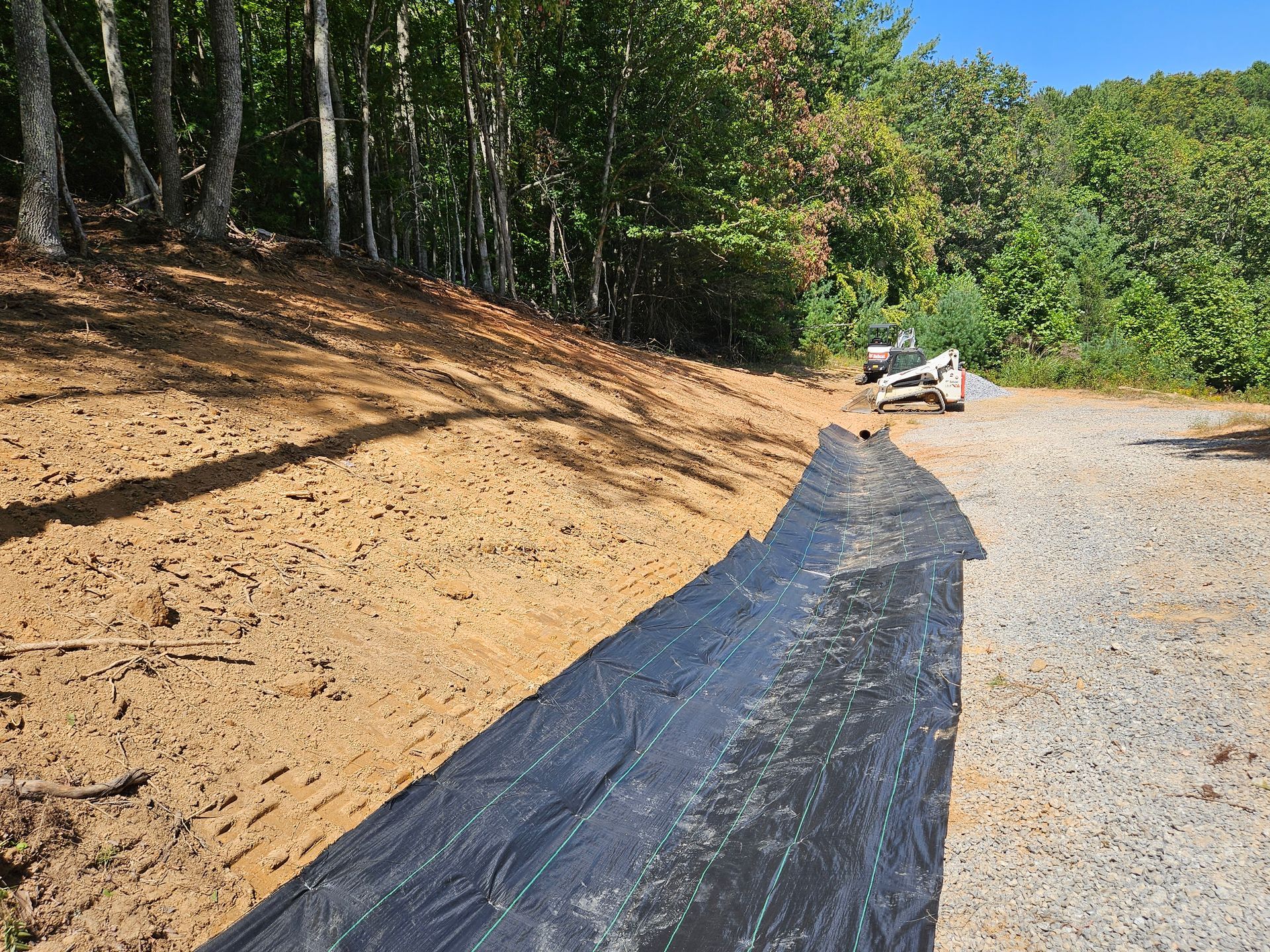 Black tarp lining a dirt slope beside a gravel road, with trees and a small vehicle in the distance