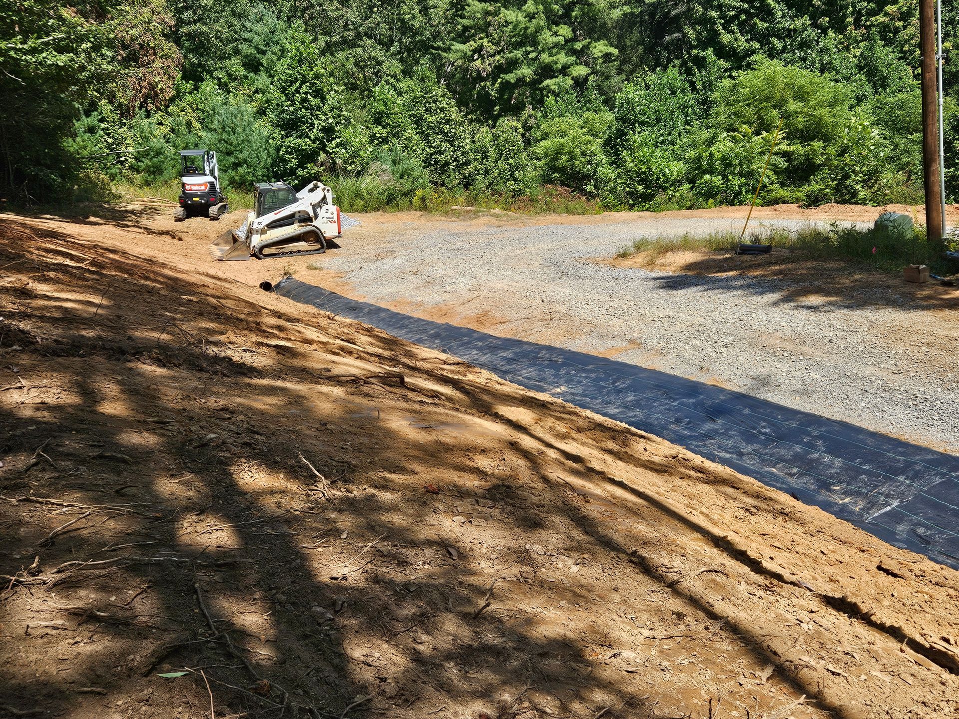 Fresh blacktop paving on a curving dirt road beside trees, with a small tractor in the background