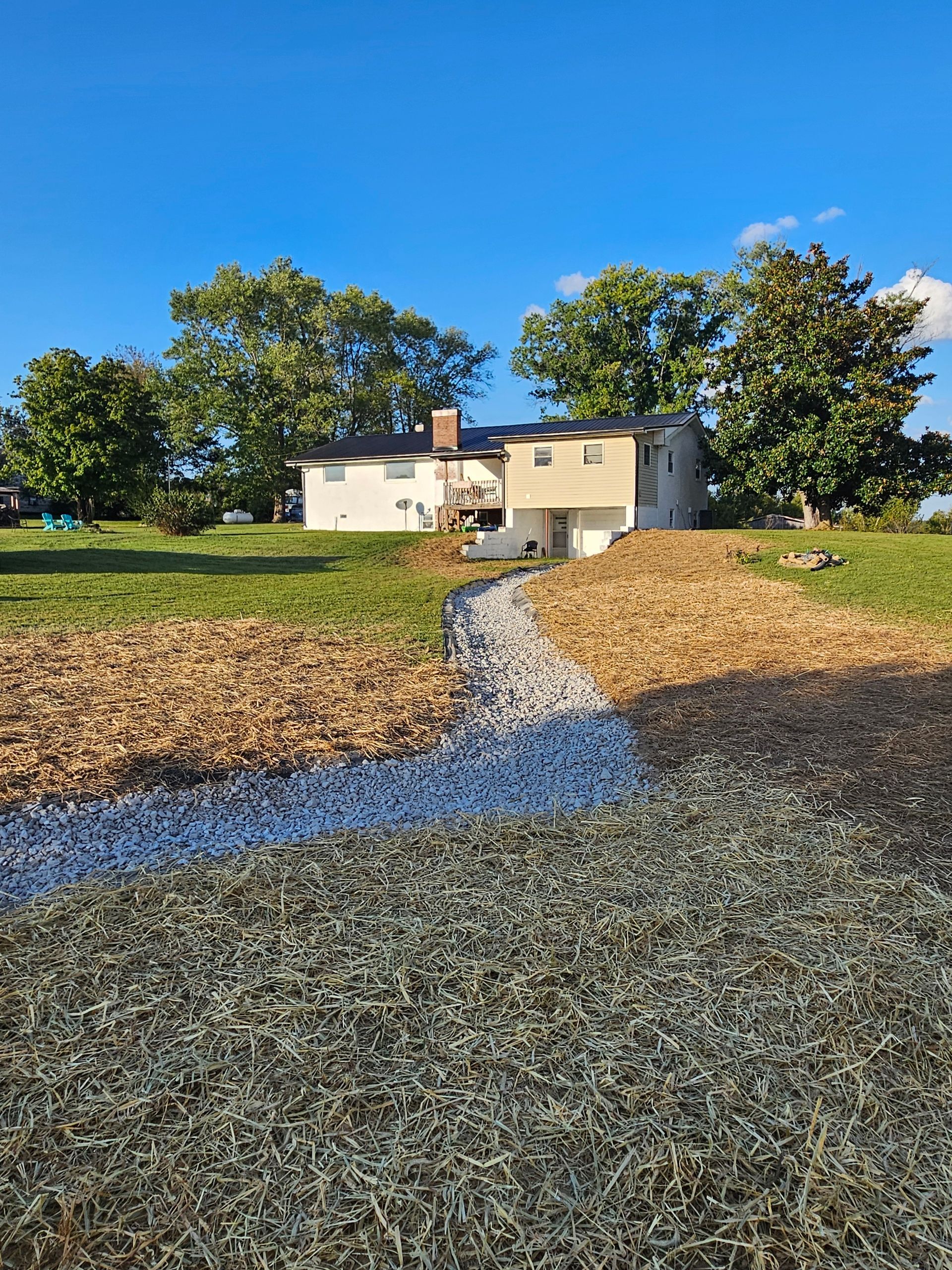 White house with chimney at the end of a gravel path, surrounded by grassy field and trees under a blue sky.