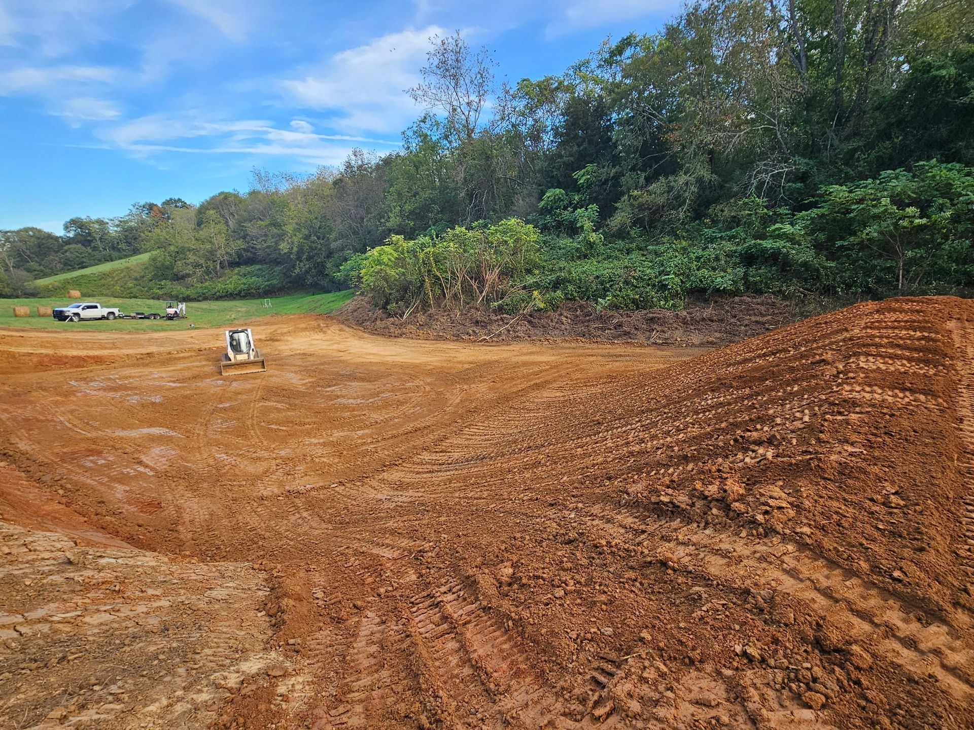 Freshly graded dirt field bordered by trees, with a small white object in the distance under a blue sky