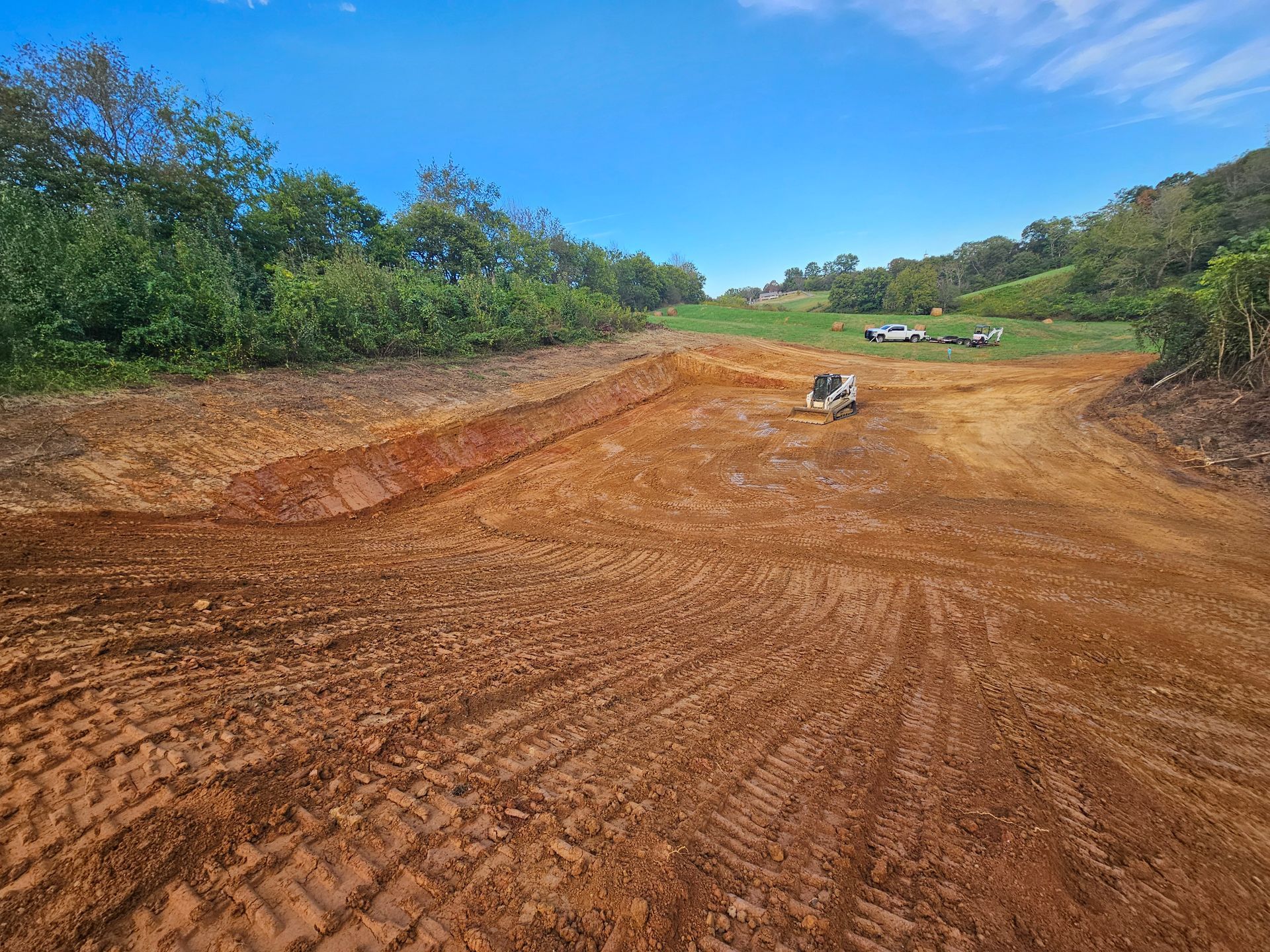 Wide dirt road under construction with a small excavator and trees lining both sides under a blue sky