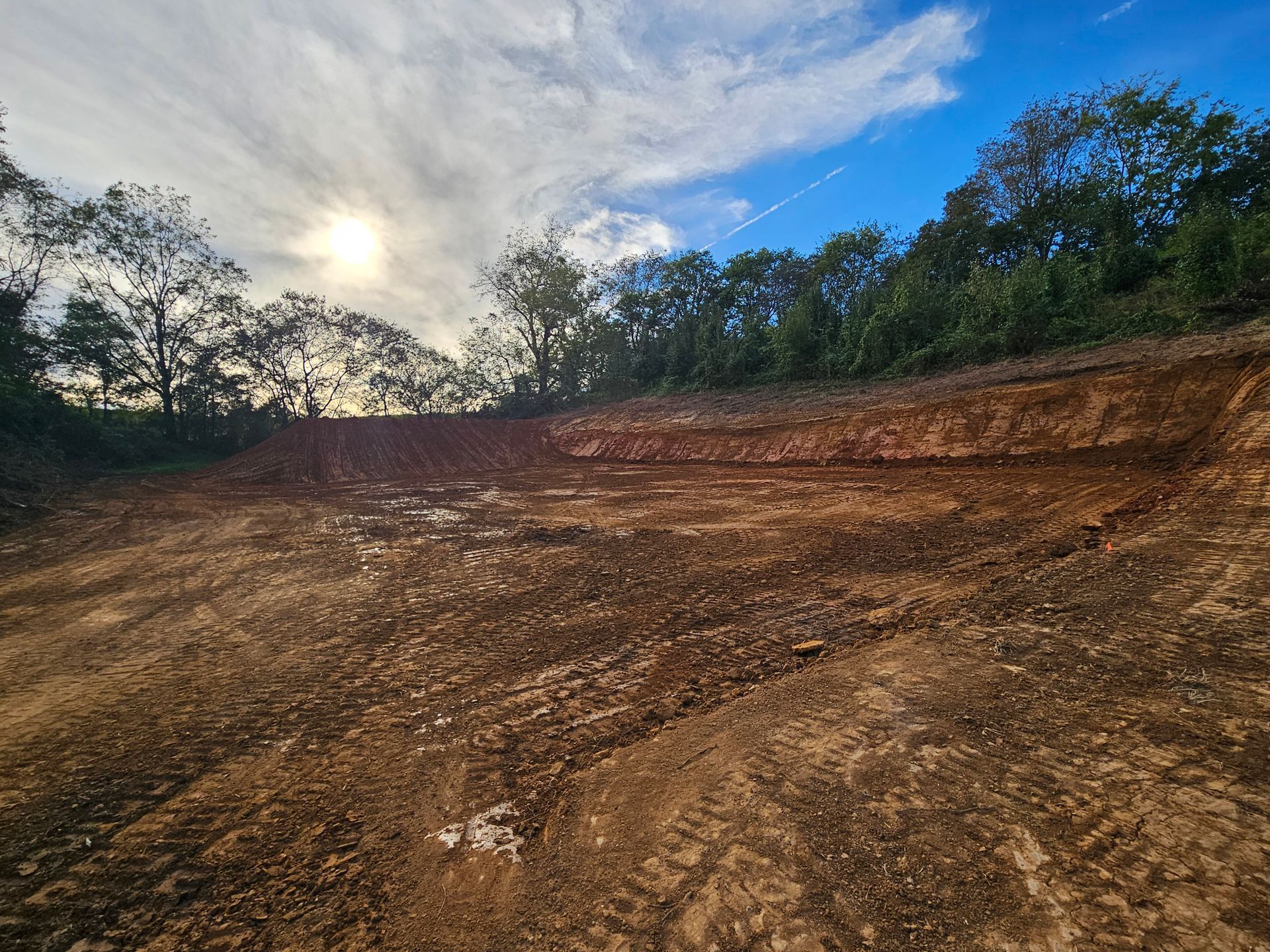 Bare dirt hillside under a cloudy sky, bordered by trees with the sun peeking through.