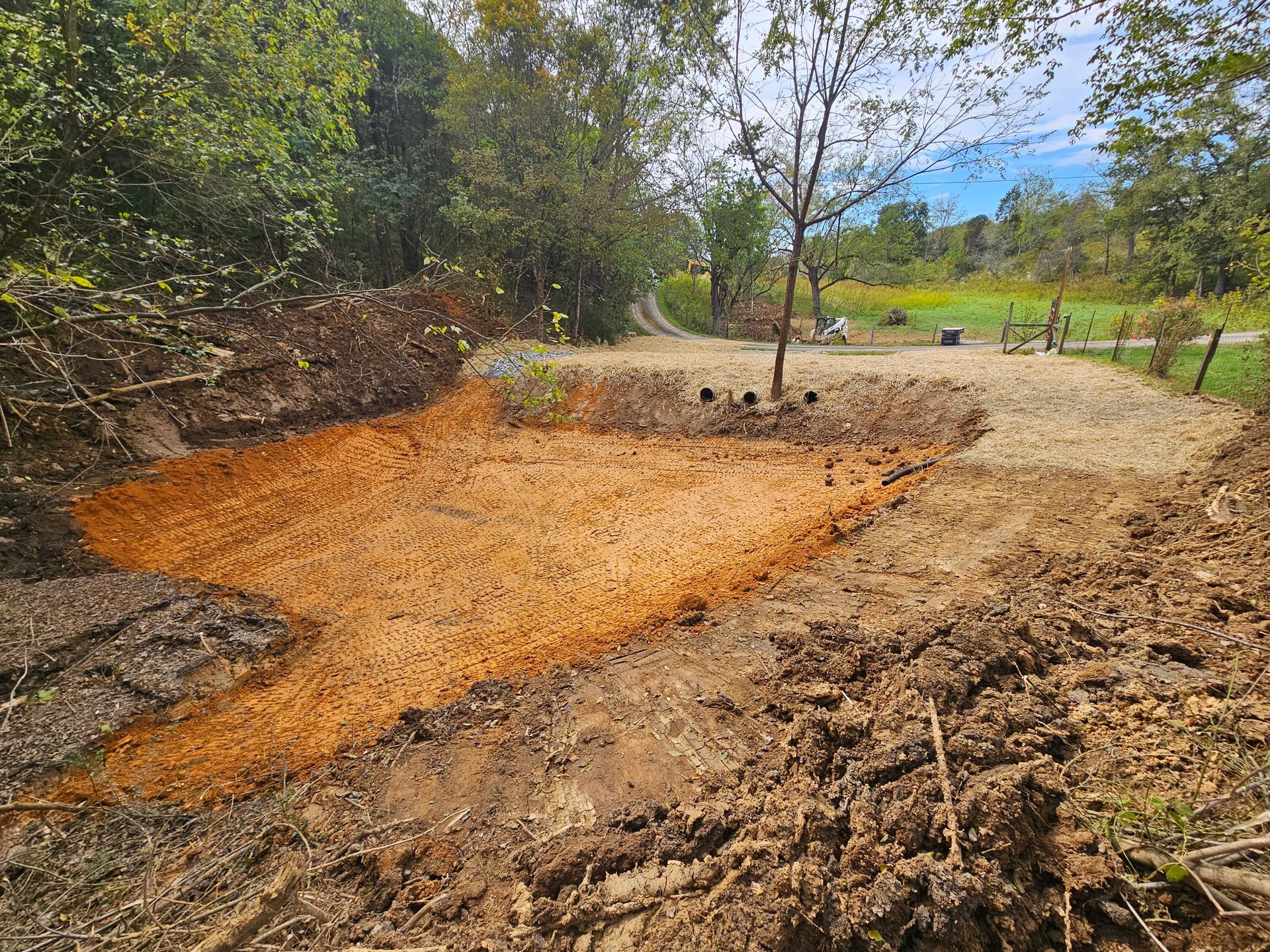 Excavated orange-brown soil pit beside a dirt road, with trees and a grassy field in the background