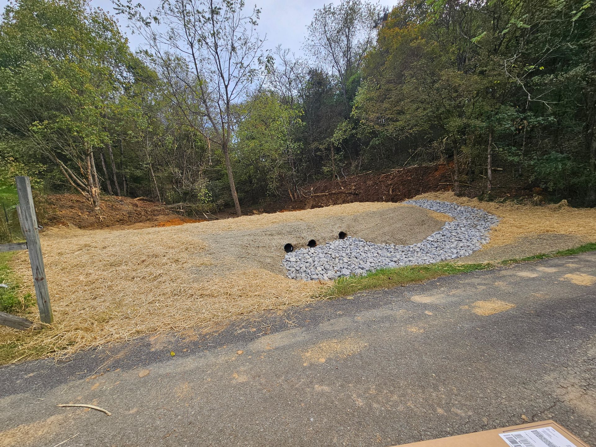 Gravel landscape with a curved stone bed, sparse plants, and trees beside a paved road.