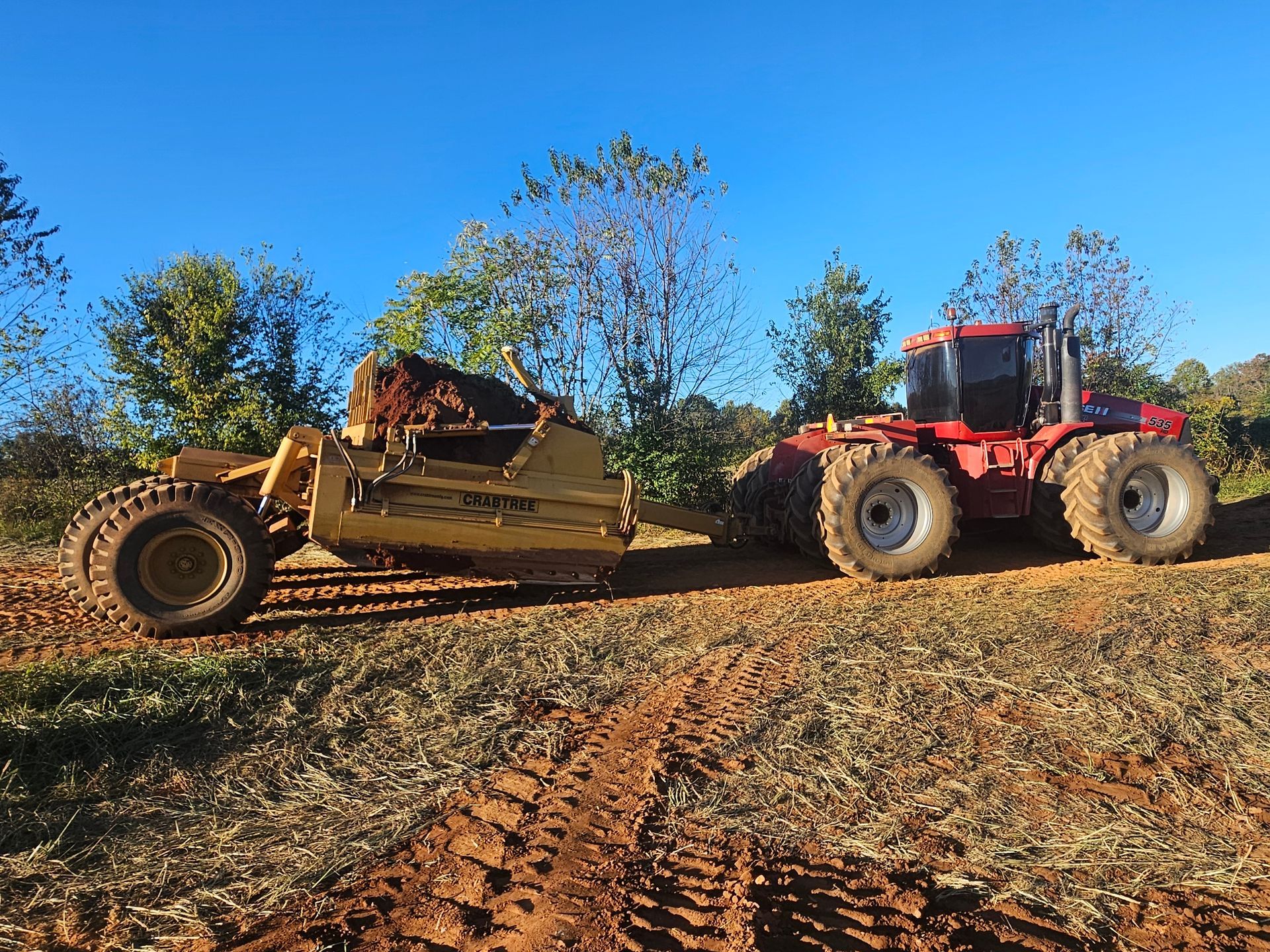 Tractor towing a dirt grader across a field under a clear blue sky