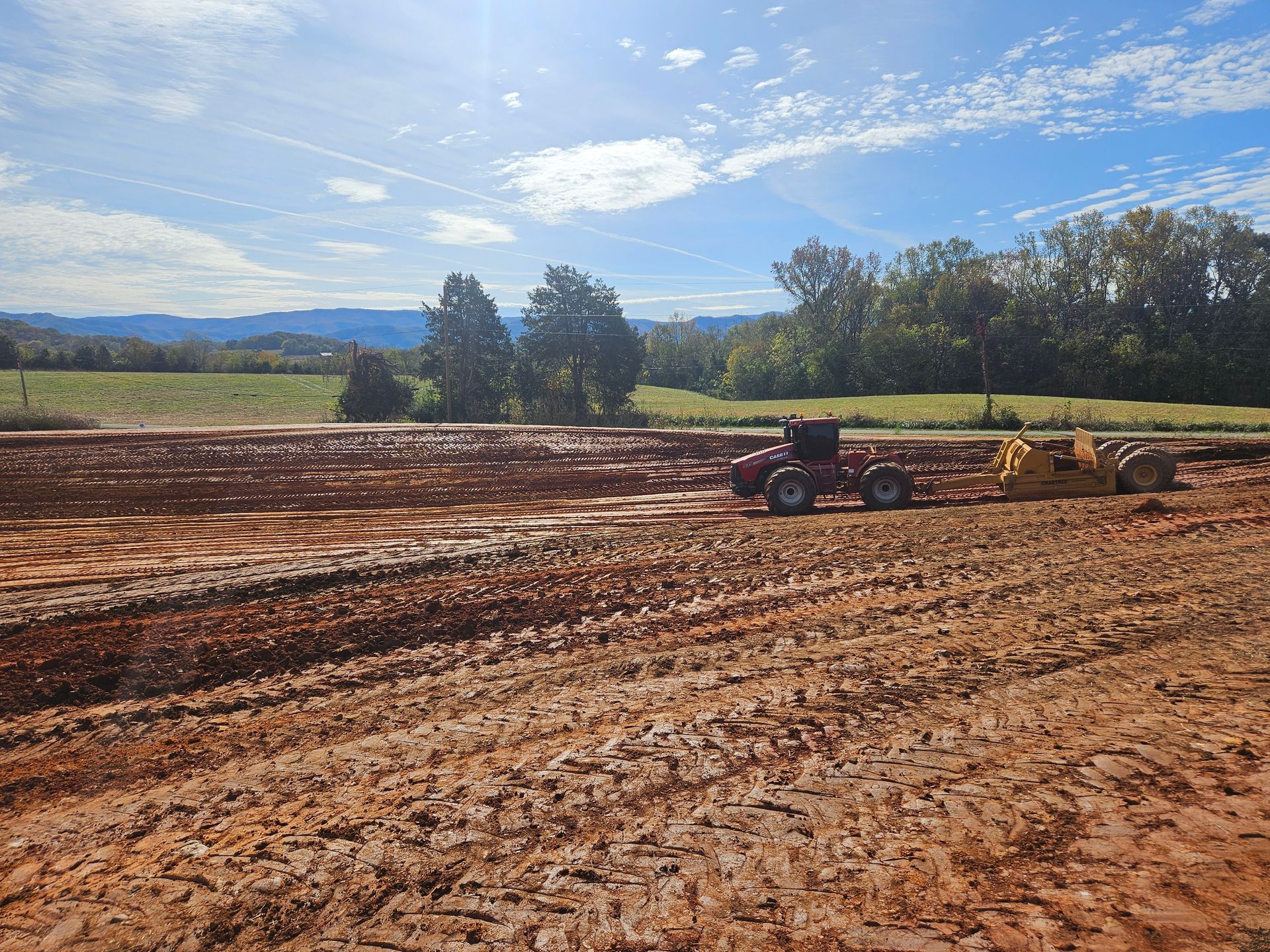 Tractor plowing a muddy field under a bright blue sky with trees and hills in the background