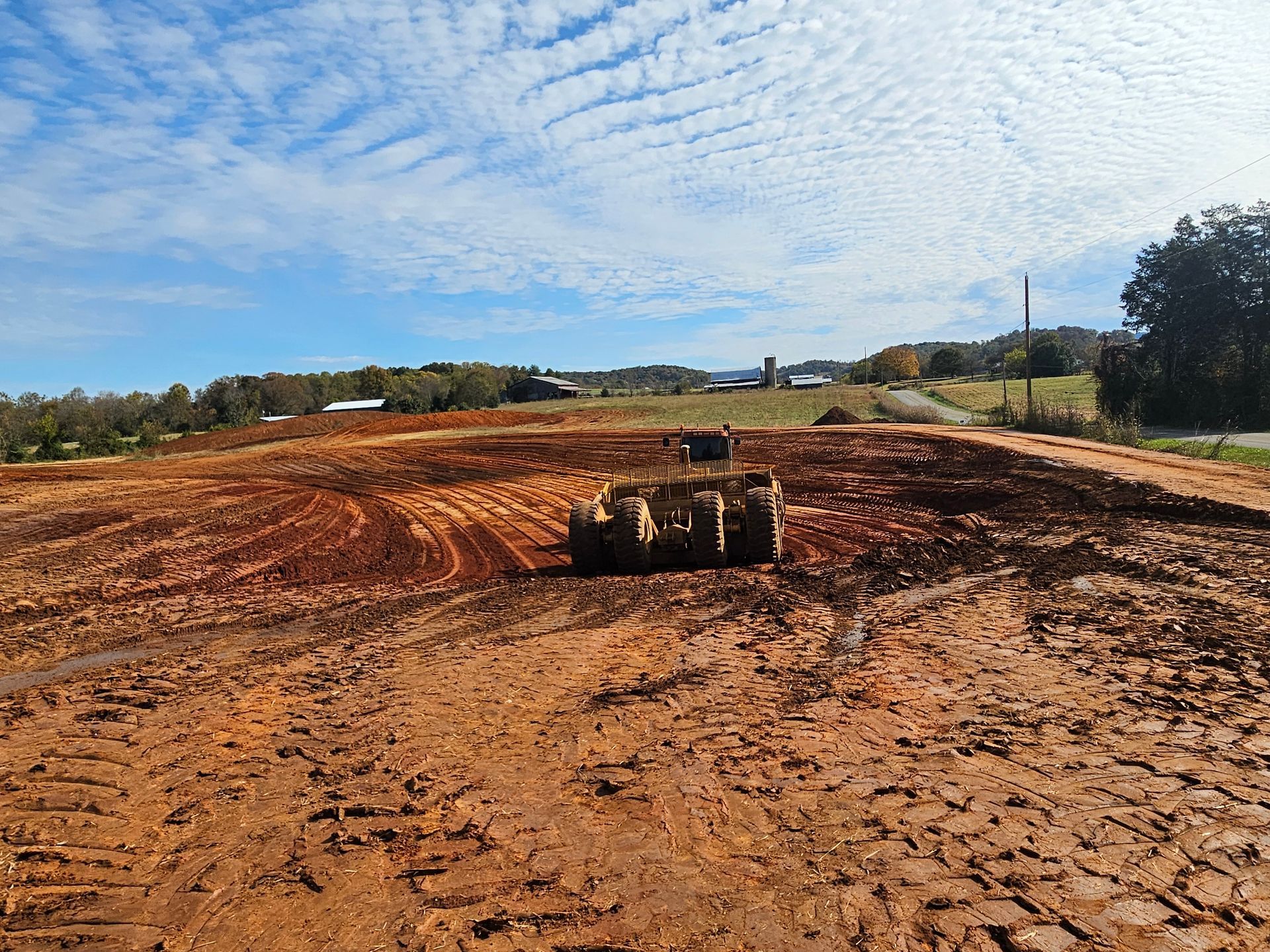 Bulldozer grading a muddy dirt field under a blue sky with scattered clouds.